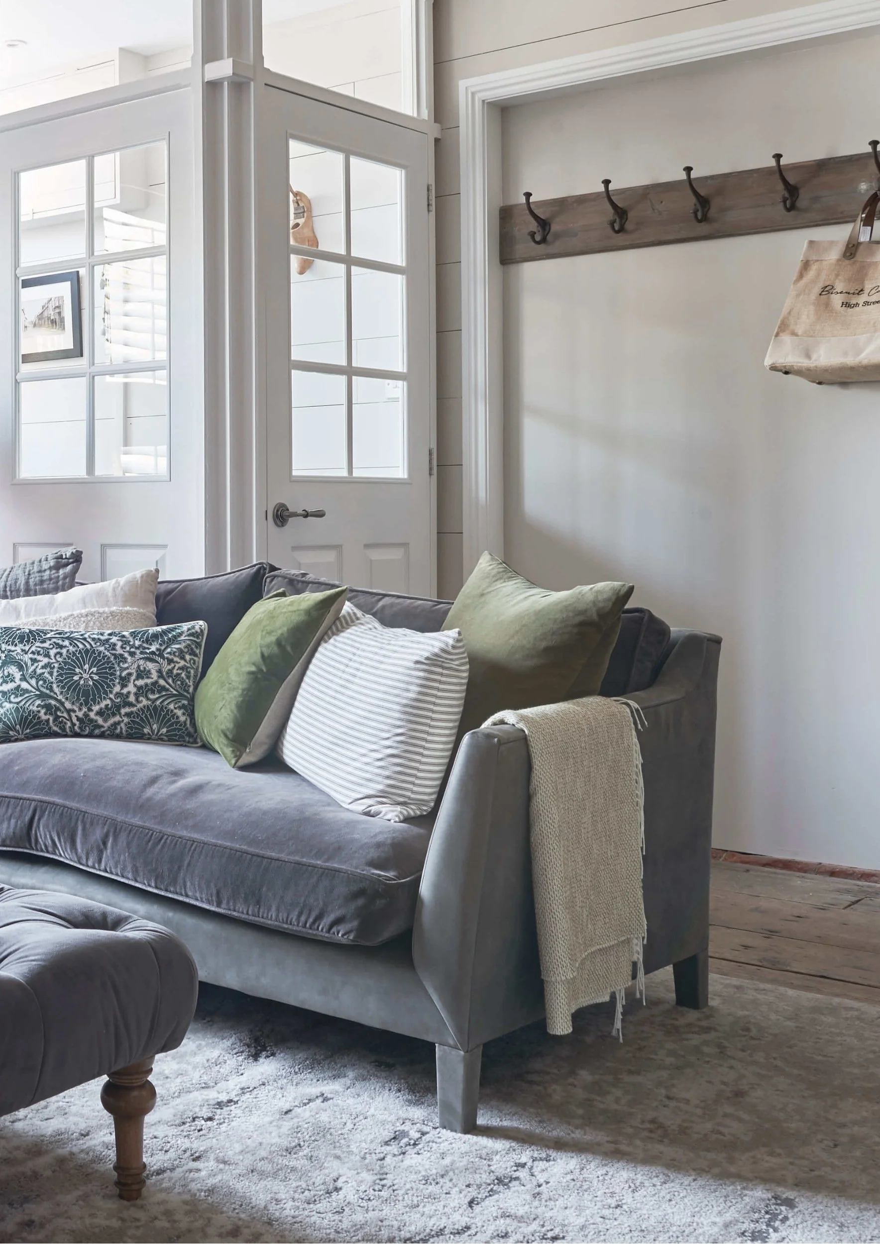 A sofa in gray/blue on top a rug with a mixture of cushions plain and patterned in front  entrance and a row of hooks