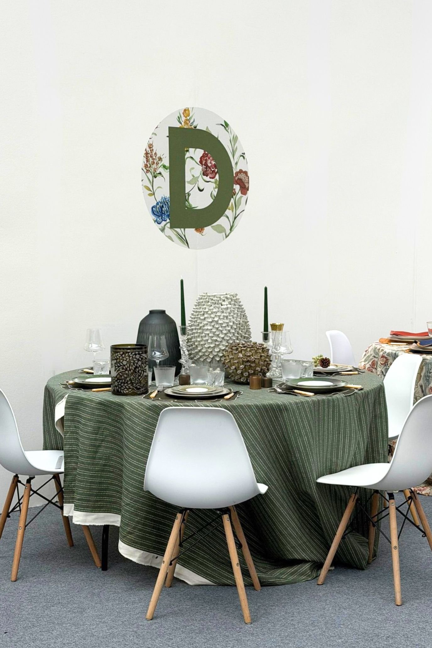 Round table draped in green fabric and styled with decorative objects, surrounded by white chairs beneath a floral Decorex D.