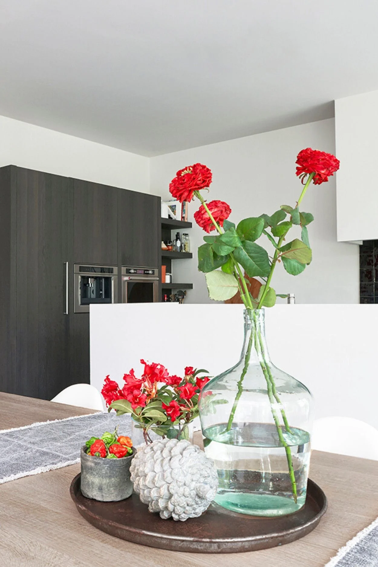 a stylish metal tray with a vintage vase with red flowers a pot with chilli peppers and decorative acorn on a wooden table with two grey  table runners and a wooden bowl with red peppers.