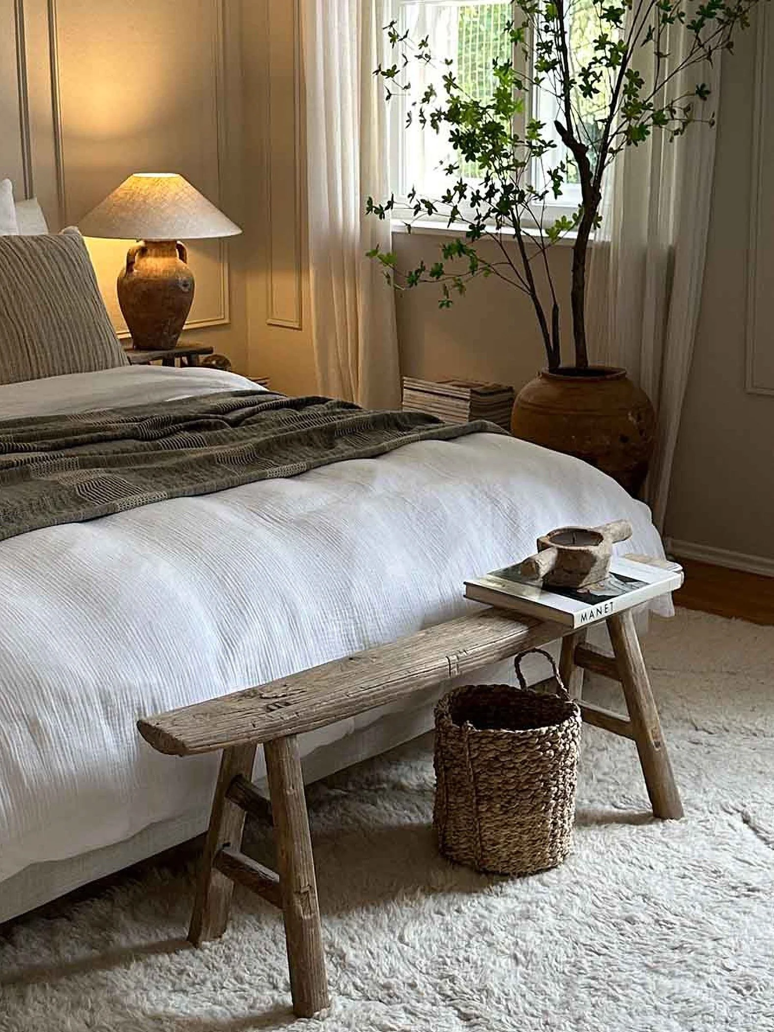 A bedroom with white linen and earthy-toned throw and cushion. There is a rustic bench at the end of the bed and a terracotta lamp and pot