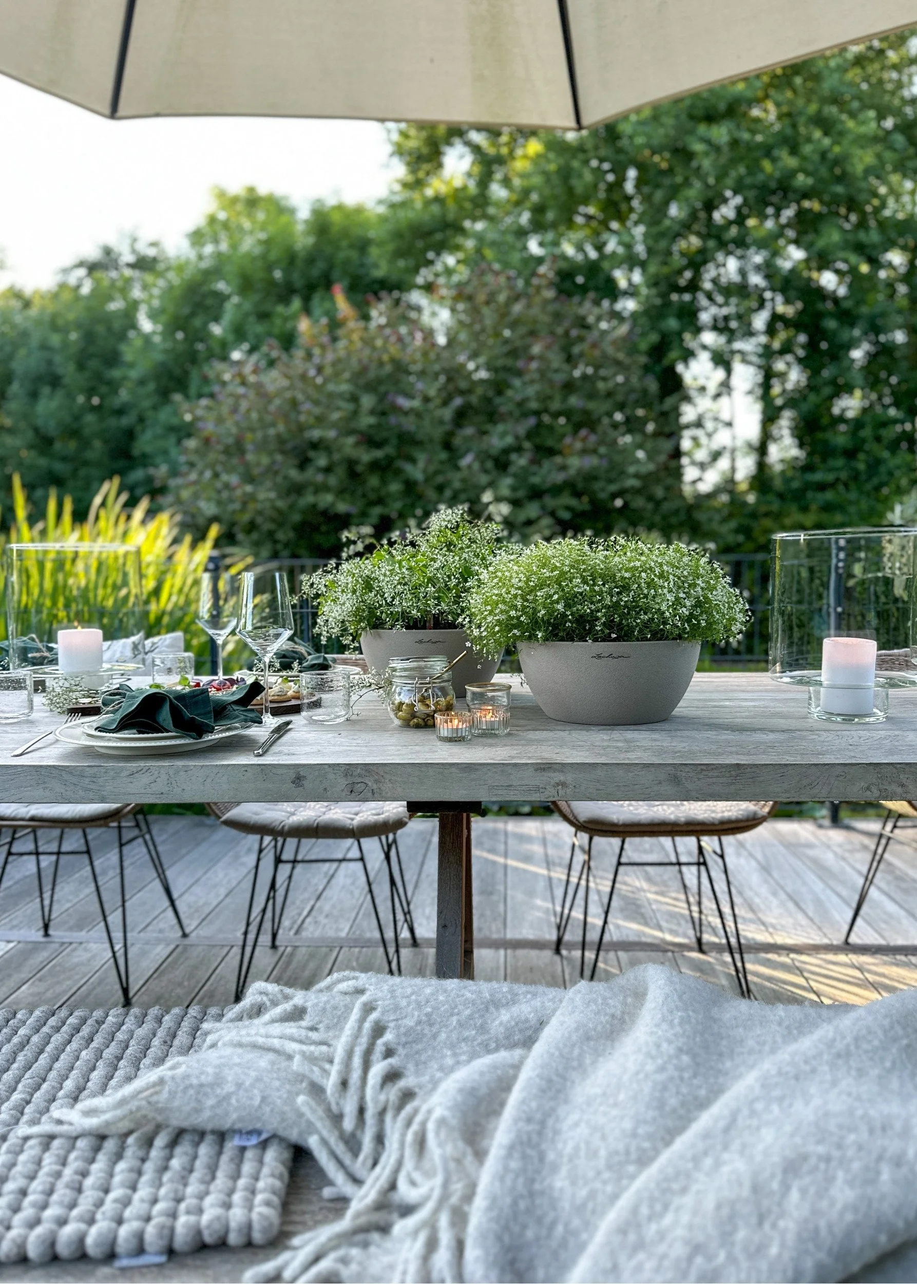 A grey outdoor table with tableware, glasses and two pots with small white flowers. There is a parasol over the table and trees in the background
