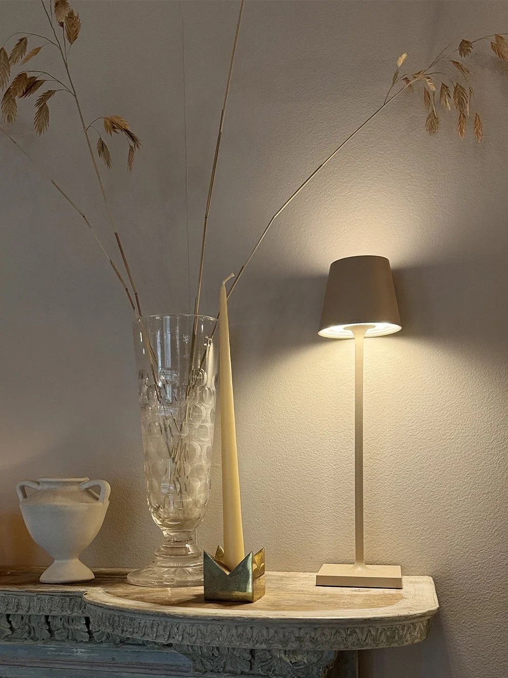 Dried grasses and leaves with a wild oat grass in a tall glass vase next to a lamp and candle stick