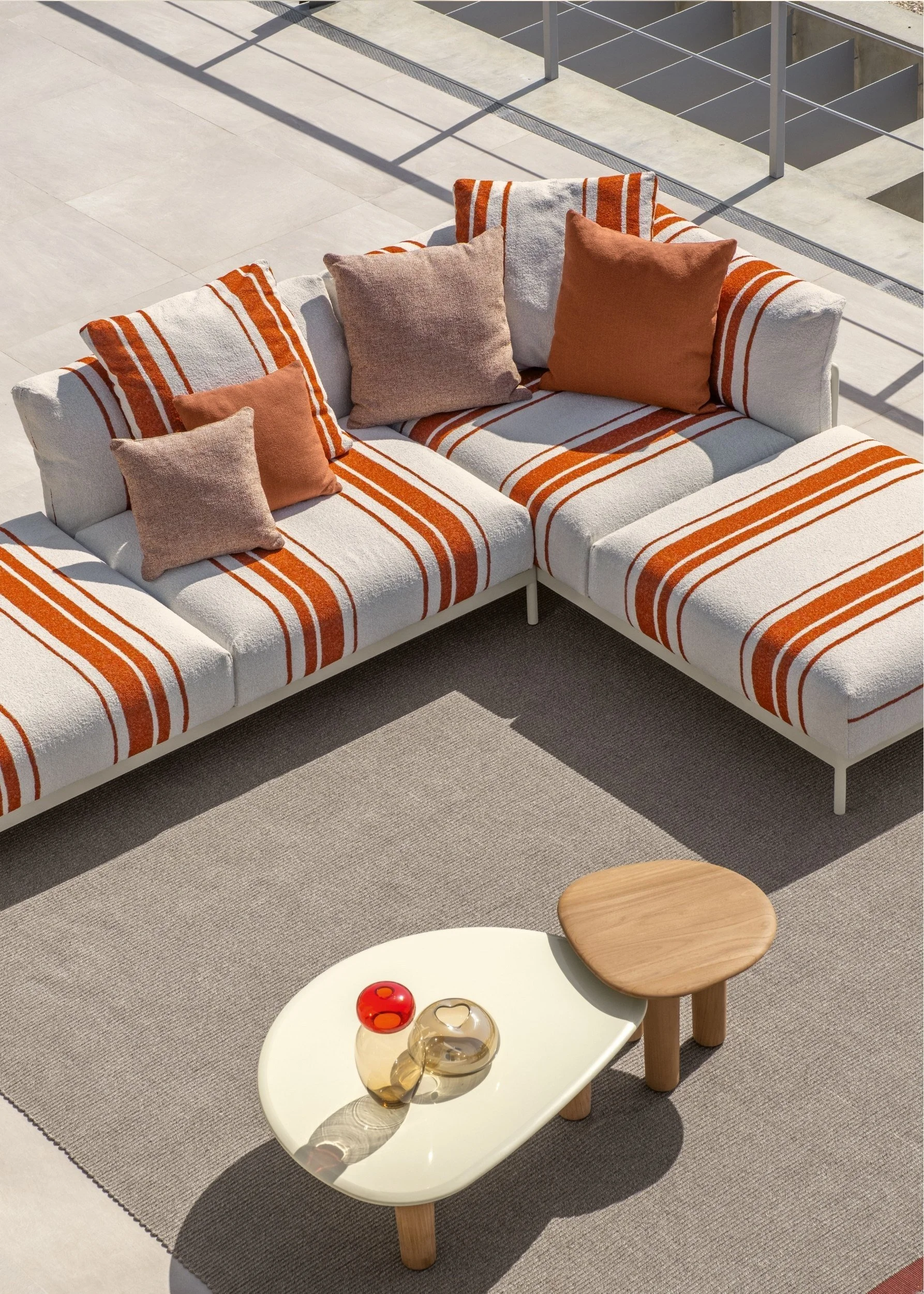 An outdoor module sofa in white with orange different sized stripes on a beige rug with outdoor coffee tables in white and wood