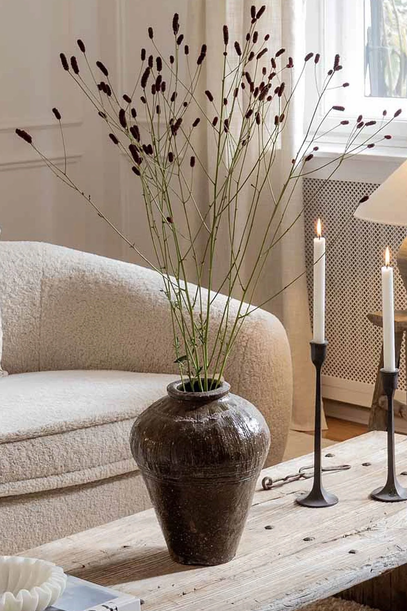 An antique vase in earthy tones displaying grasses on a rustic table next to two iron candle sticks
