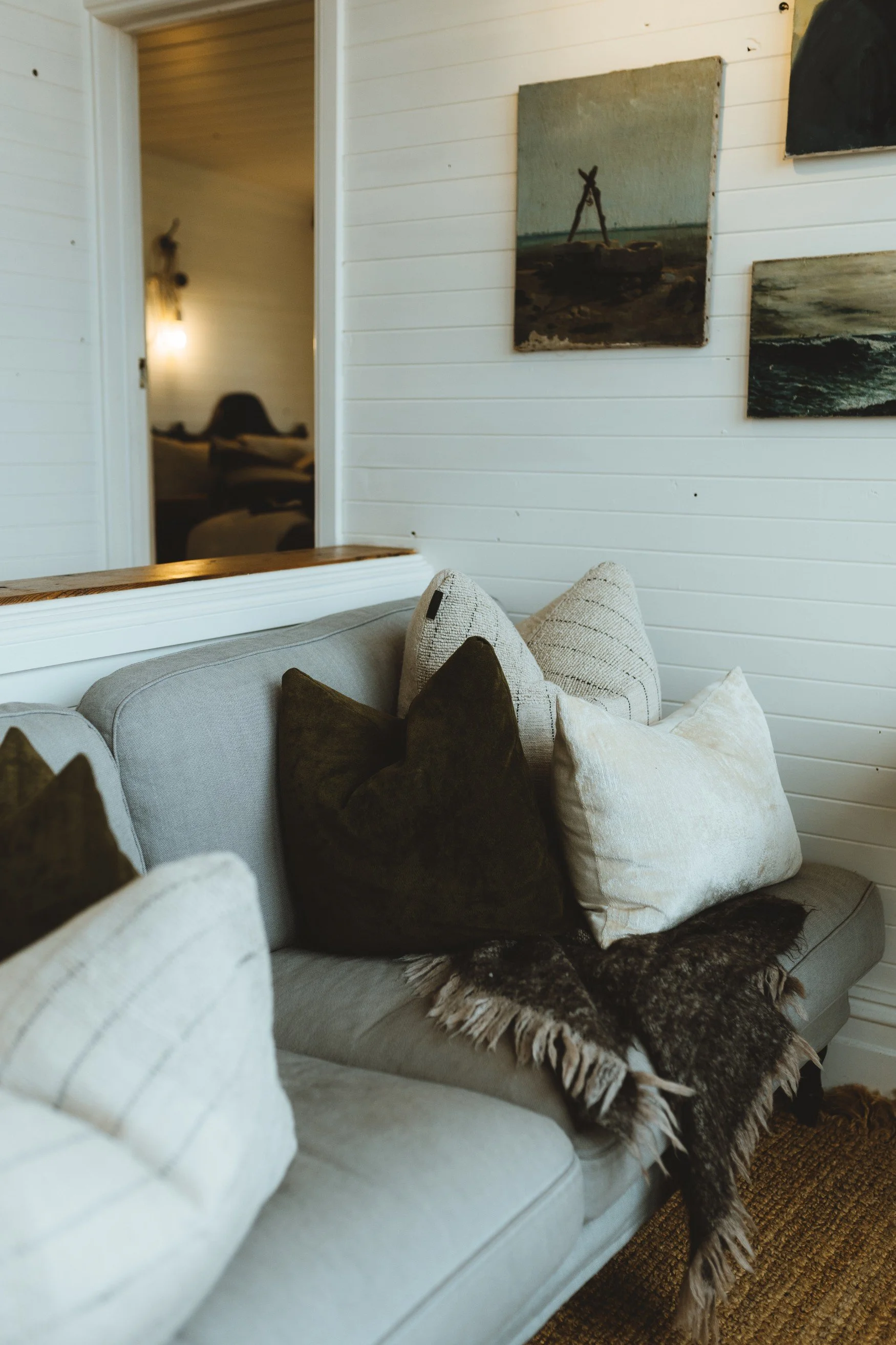 A sofa styled with cream and brown cushions with a brown wool throw and old paintings on the wall.