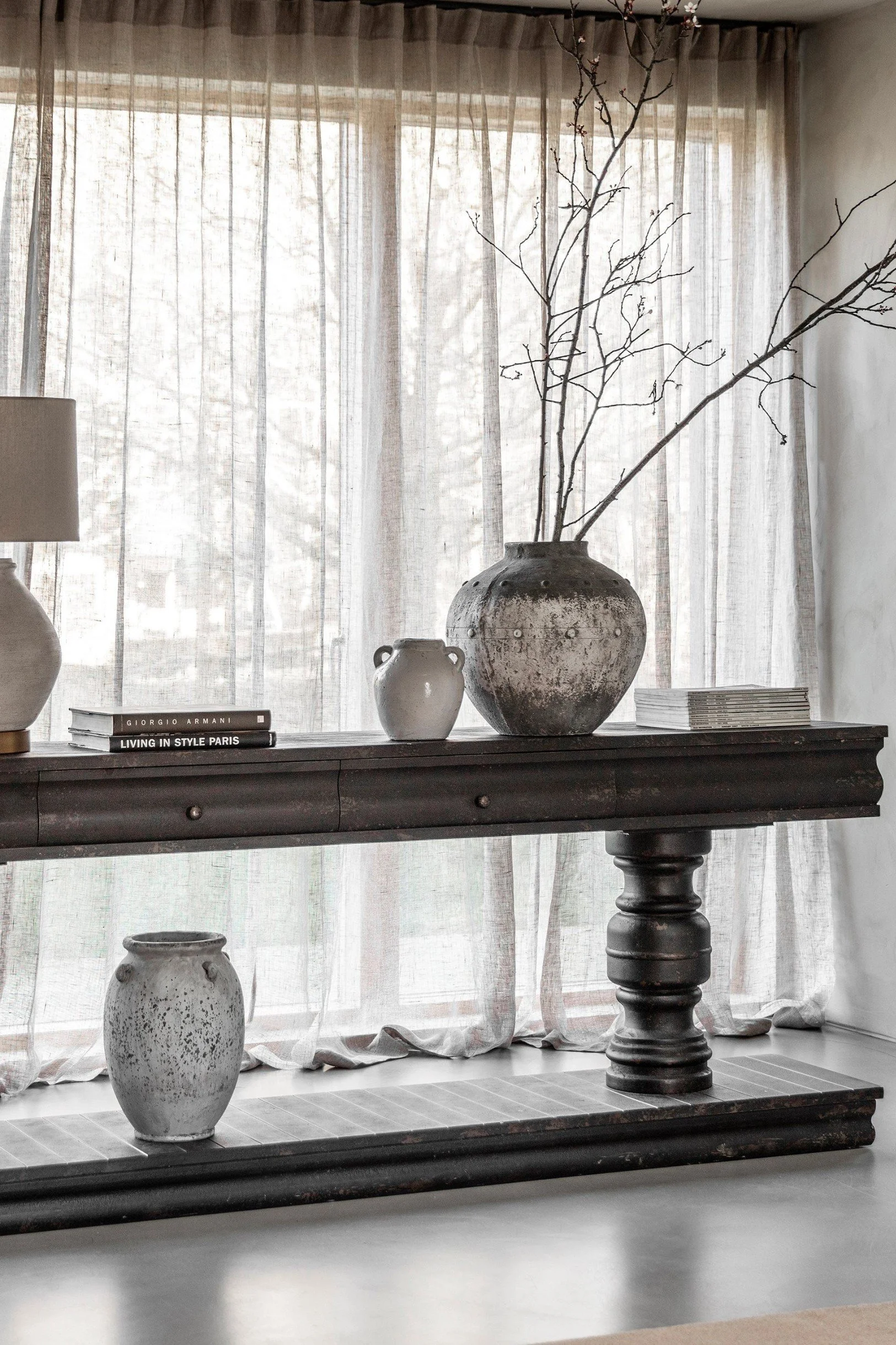 A wooden console table with a large vessel with branches next to a small white pot with piles of magazines and books either side and a lamp on the left