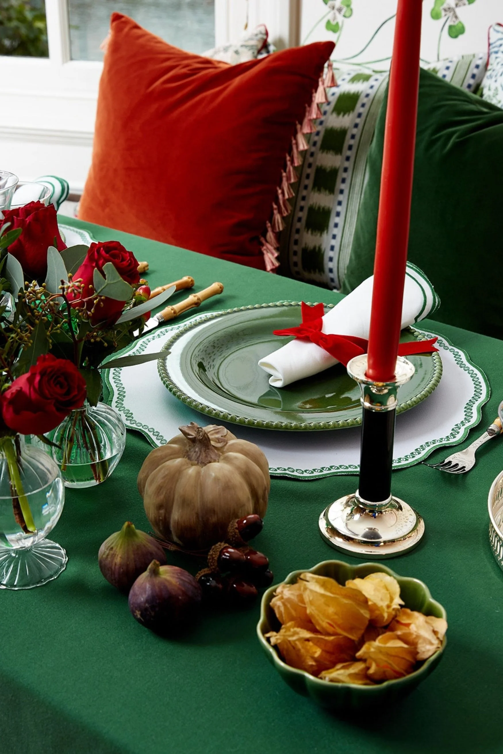 Silver candle holder with red candle on a green table cloth with red roses in vases and a red and green cushion behind the table