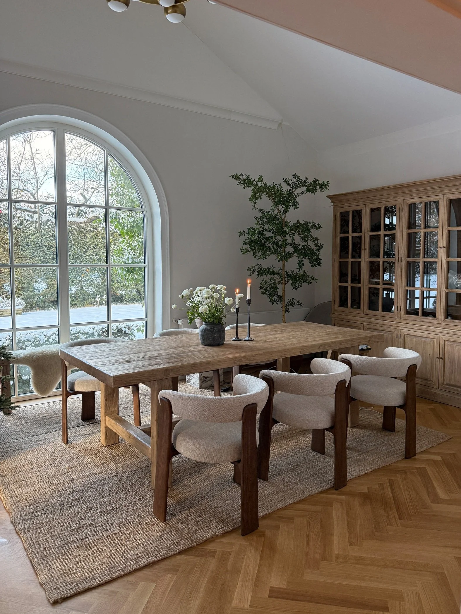 A dining room with an arched window a large wooden table, cream and wood modern chairs and a classic wood and glass cabinet
