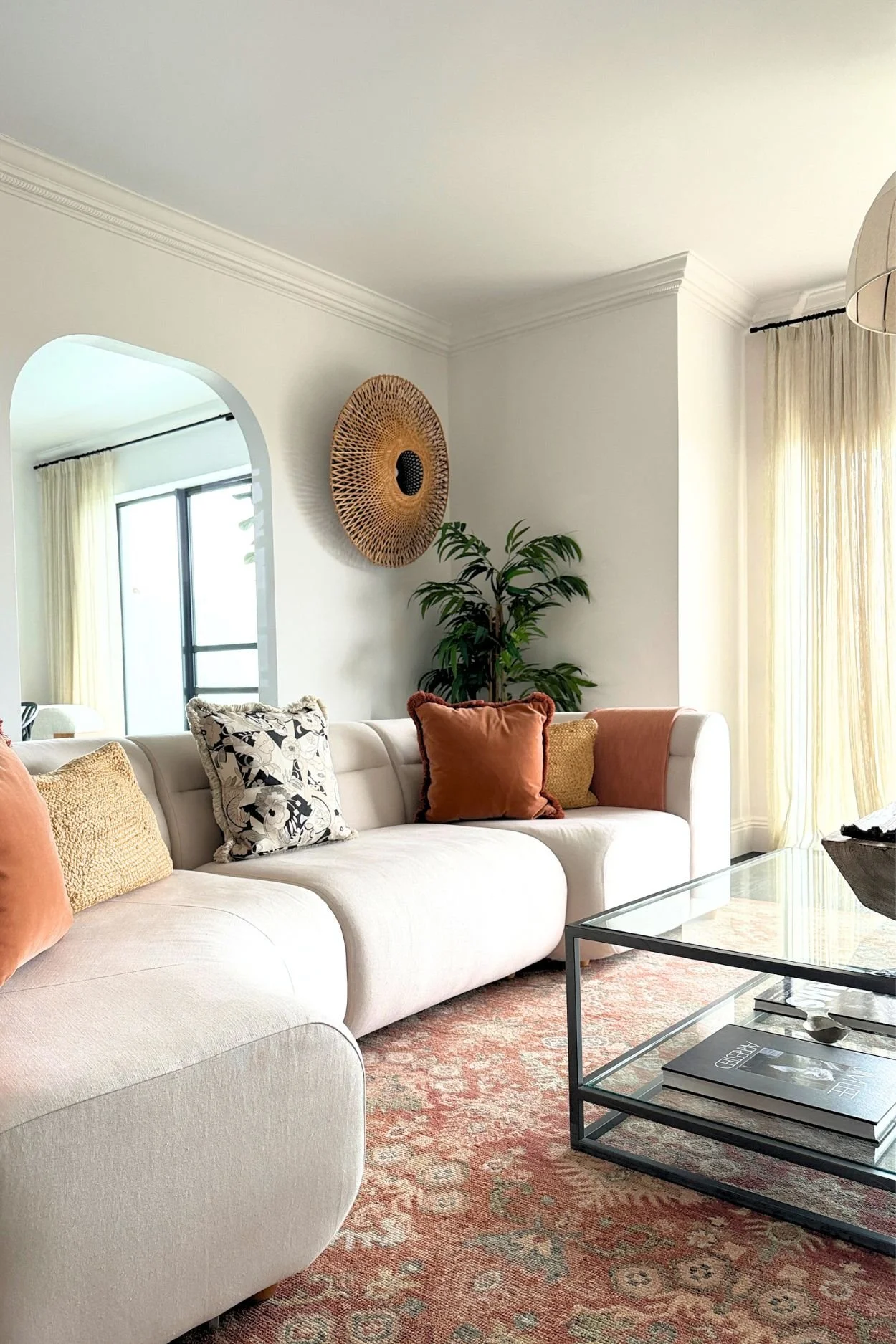 An off-white sofa with terracotta toned cushions and rug infront of a plant and round rattan wall light