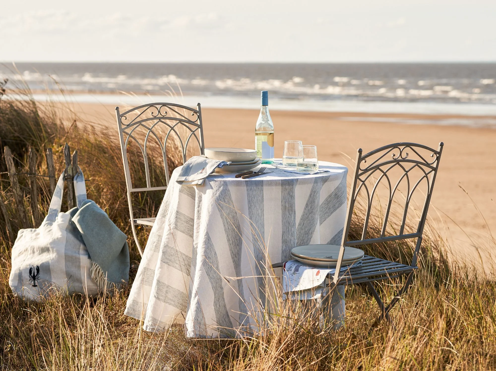 An outdoor table and chairs in long grasses next to a beach styled with a striped linen cloth and tableware