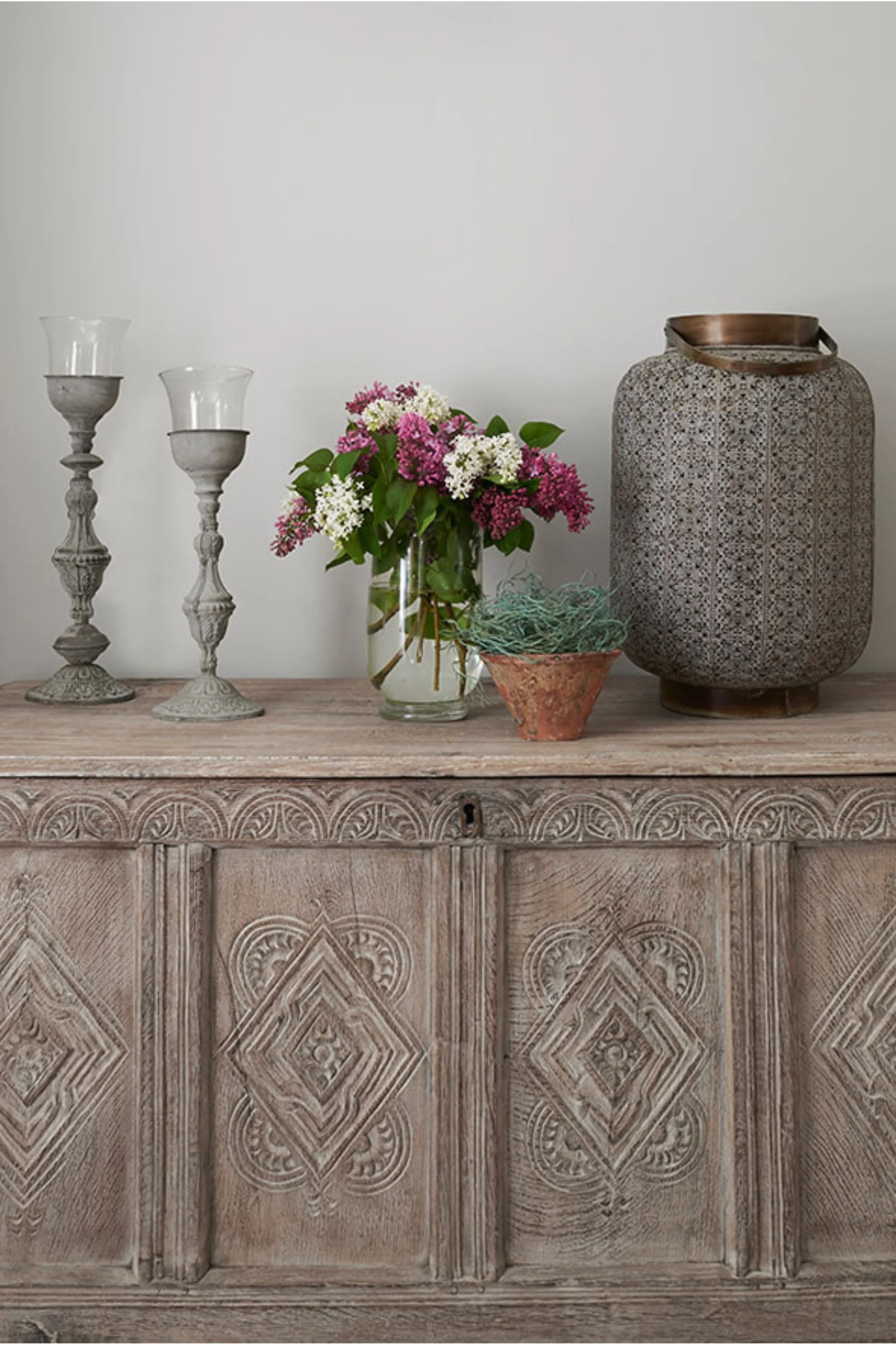 A Moroccan trunk styled with a large lantern, two candle holders, a vase filled with lilac and a potted plant