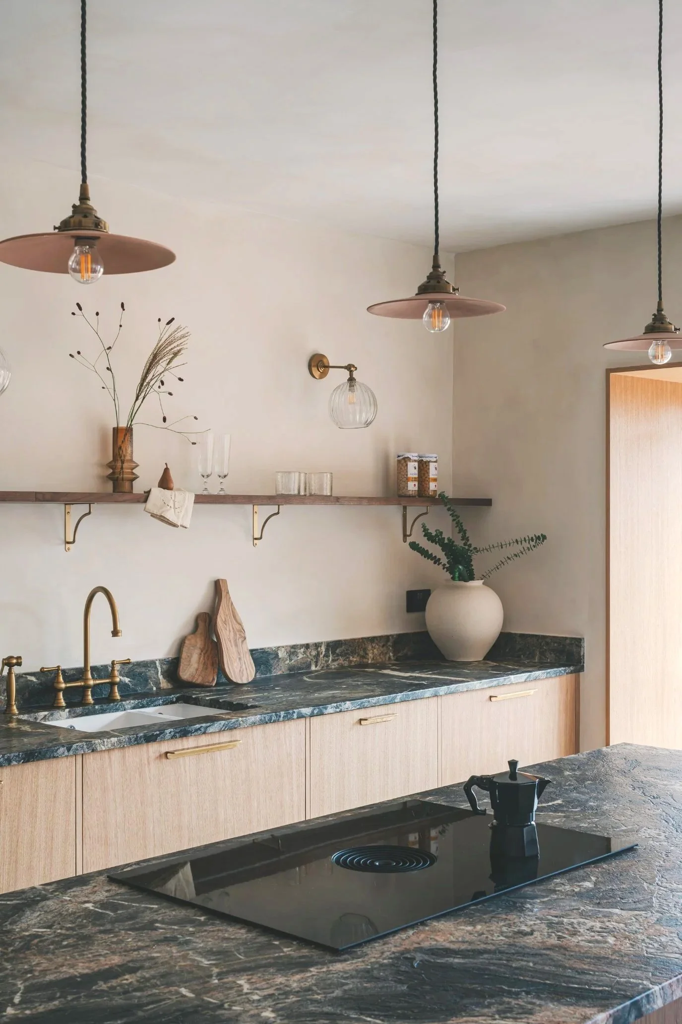 A stash kitchen with light wood cabinetry and dark marble countertops with a single shelf above and items like glassware and jars on display