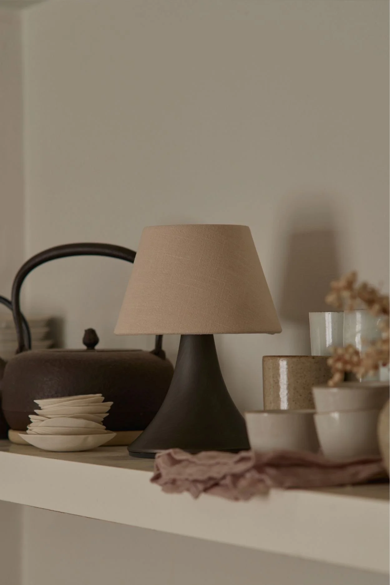 A bronze and linen lamp next to two vintage tea pots and stacked earthenware cups