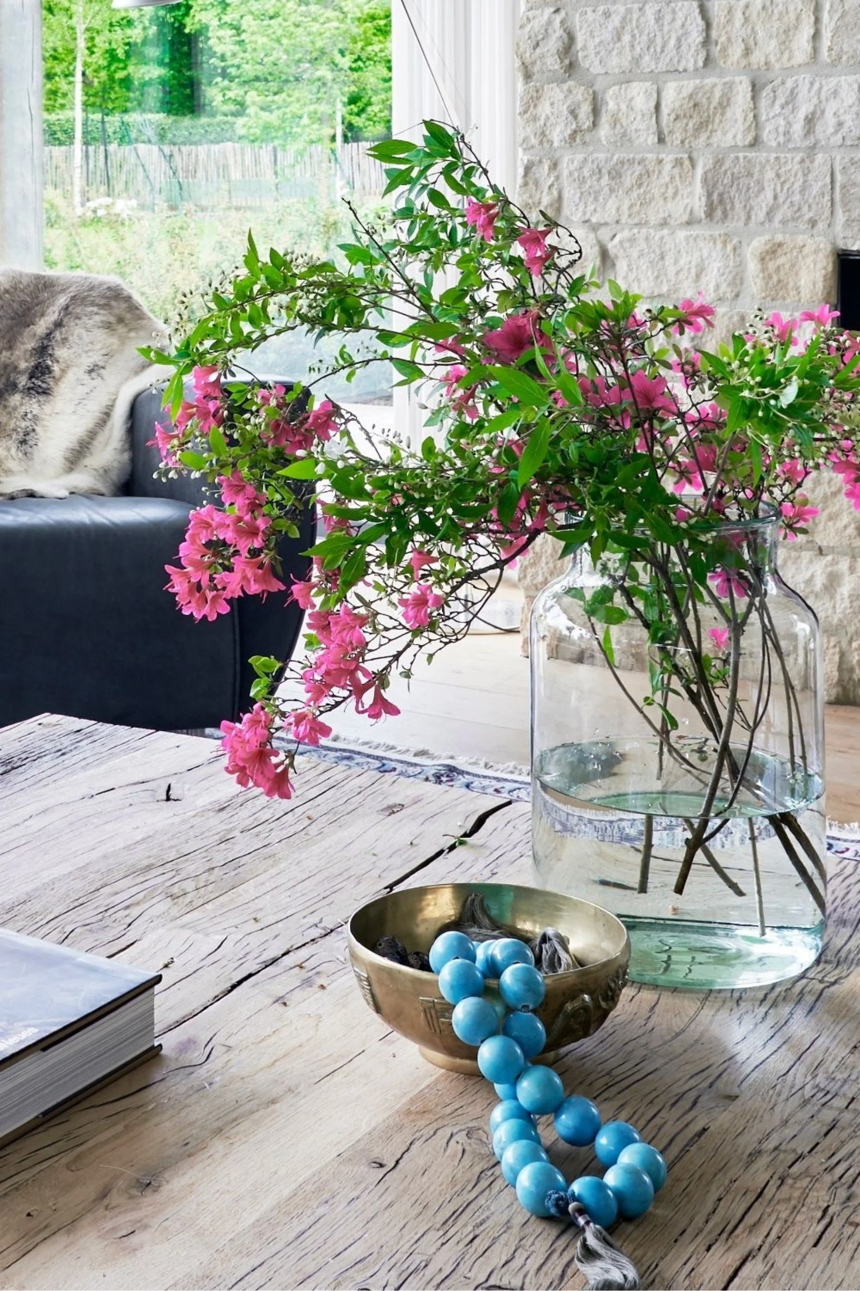 a rustic table with a vase of  pink azaleas and a brass bowl with aqua toned beads