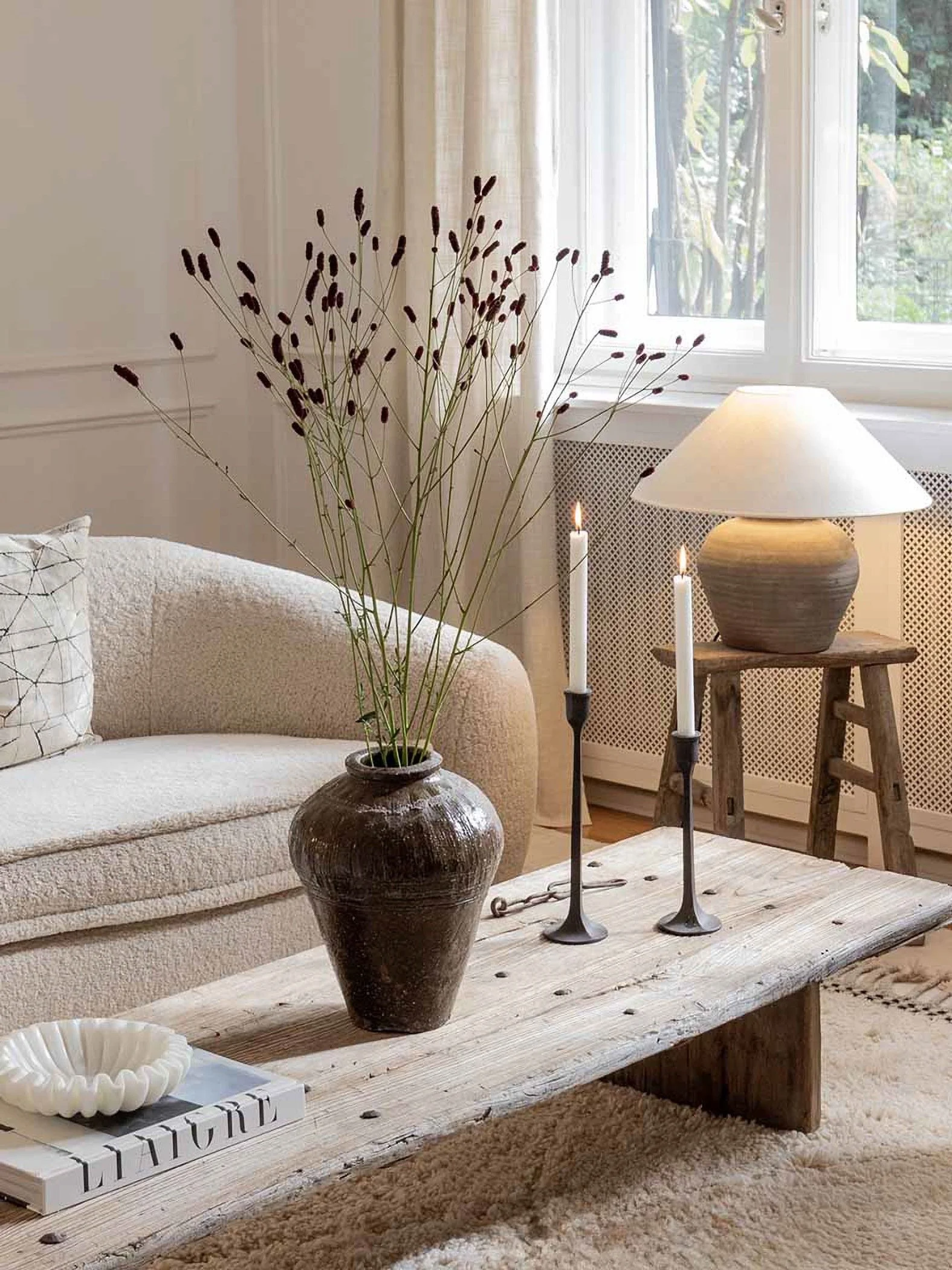 An antique vase next to two candle sticks on a wood table infront of a cream sofa which is next to a rustic stall with a lamp on it