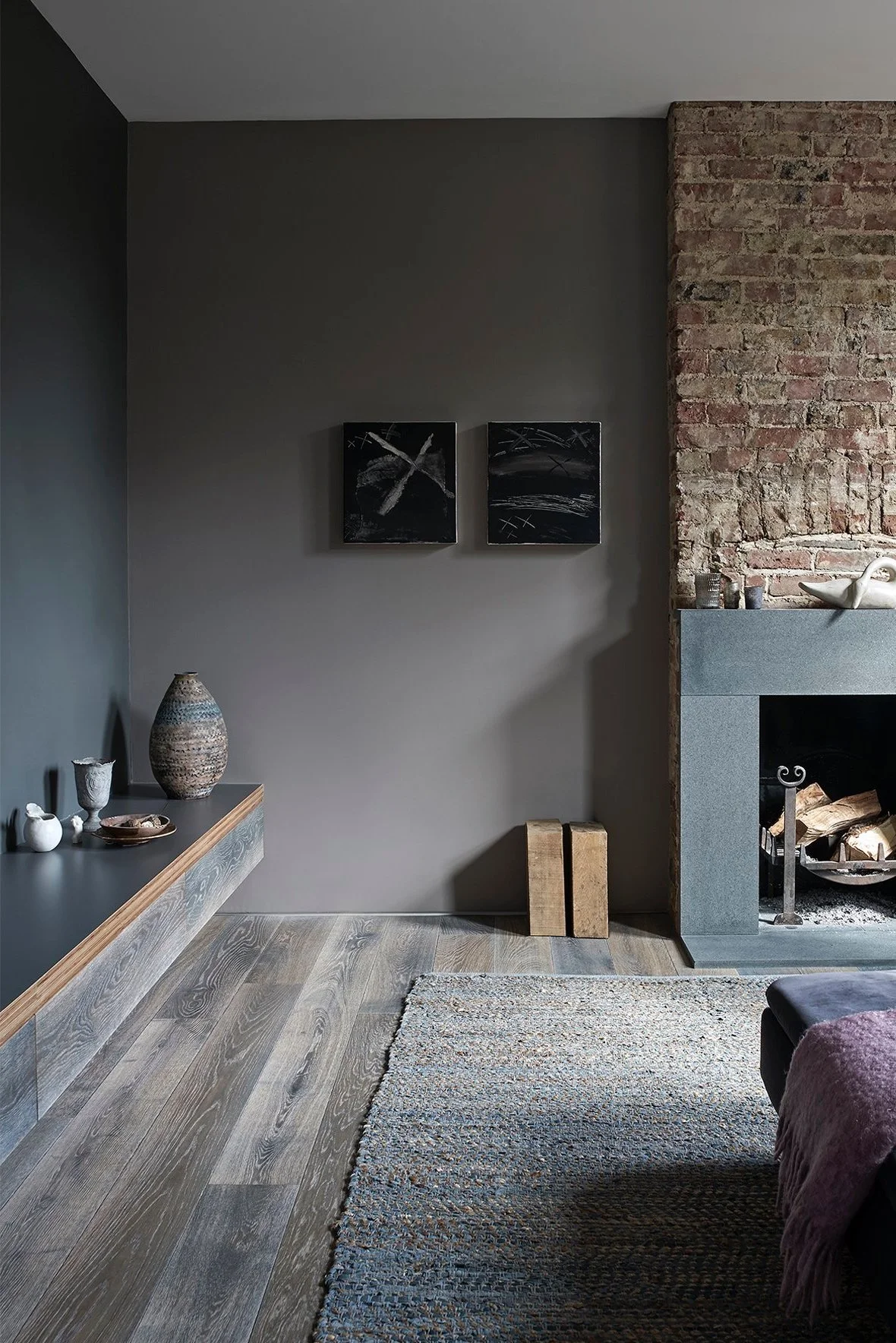 A dark grey living room with a hearth, wooden floor, rug and a low level shelf with pots on it
