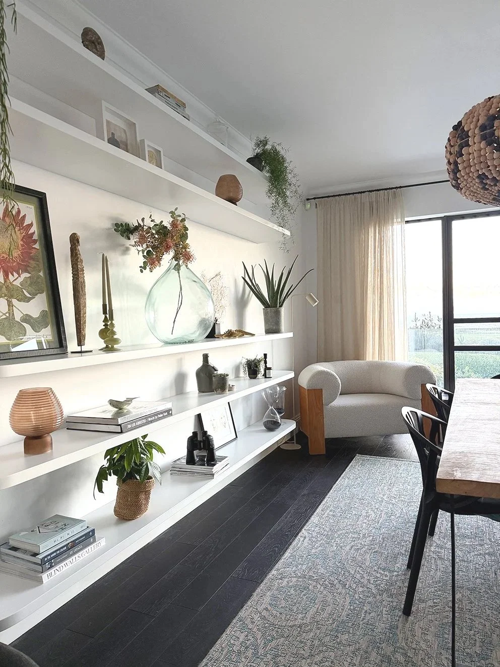 A ding area with white floating shelves and arm chair, a wooden table and beaded light