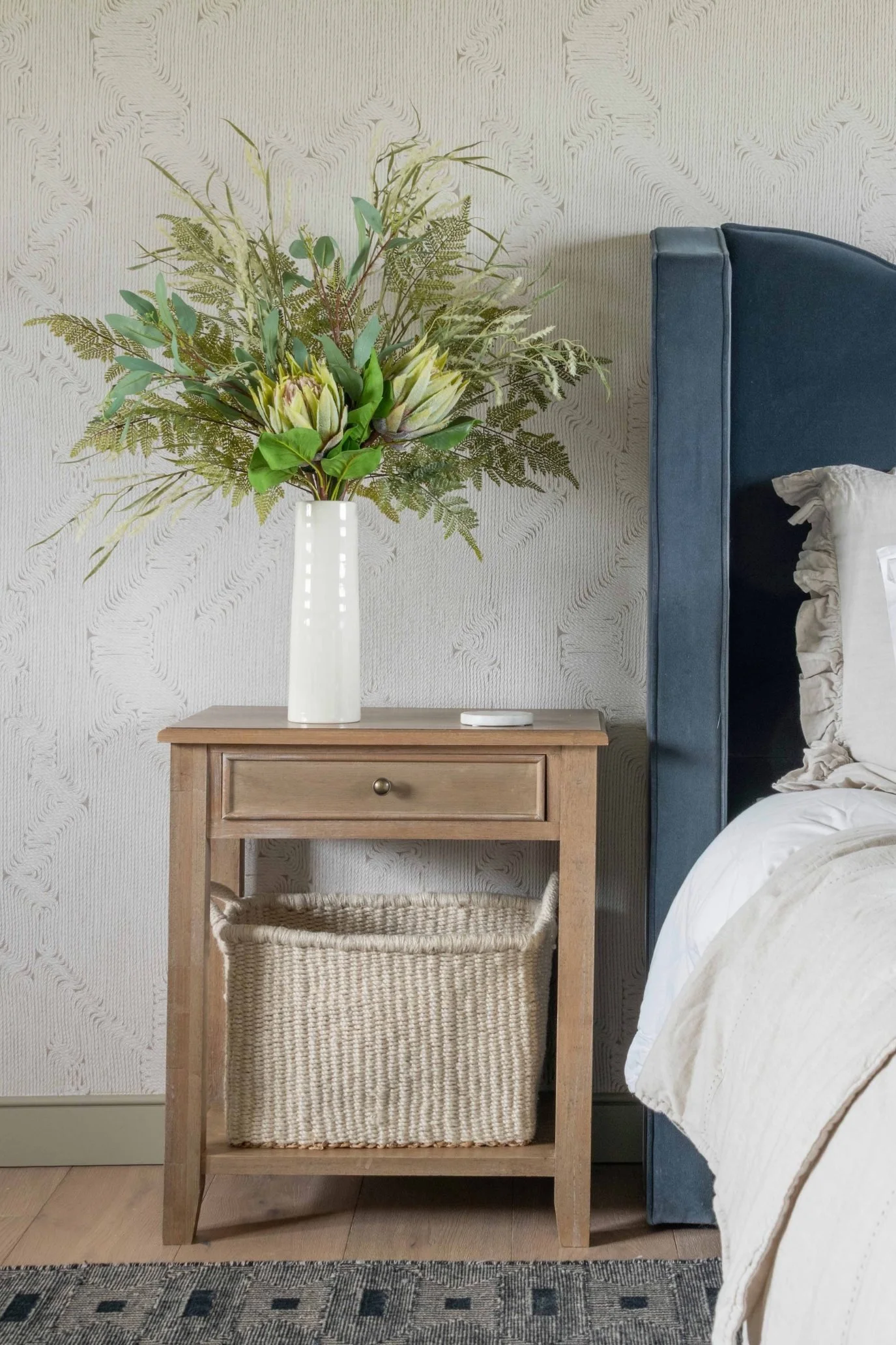 Faux proteas and foliage arranged in a tall white vase on a bedside table.