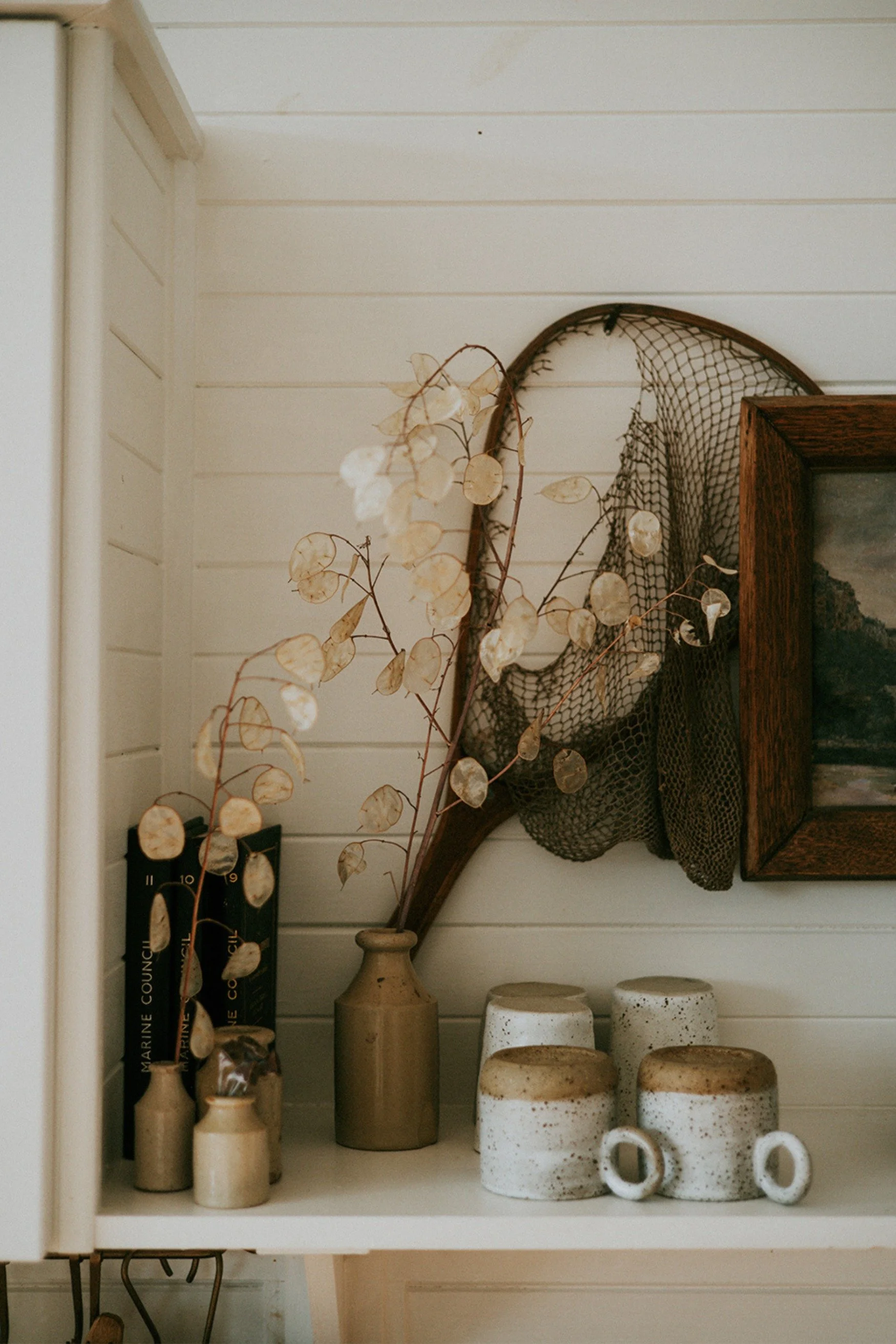 A kitchen shelf with four stoneware cups upsidedown next to small pots with seedpods and a fishing net