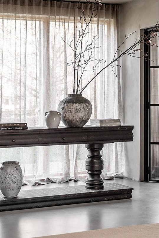 A wooden console table with a large vessel with branches next to a small white pot with piles of magazines and books either side and a lamp on the left