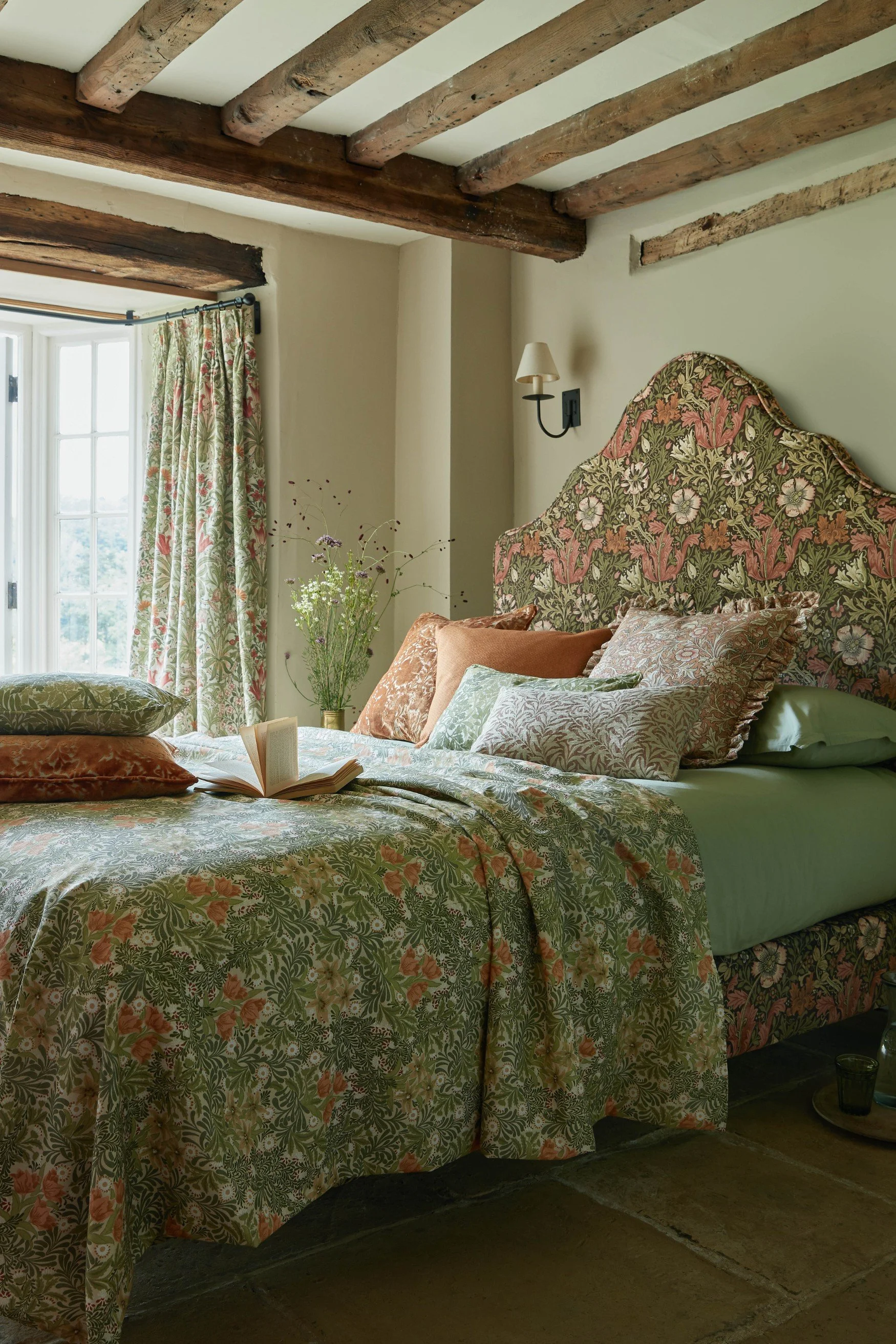 A bed with a Rococo style fabric covered headbourd in a cottage style bedroom with low beamed ceilings
