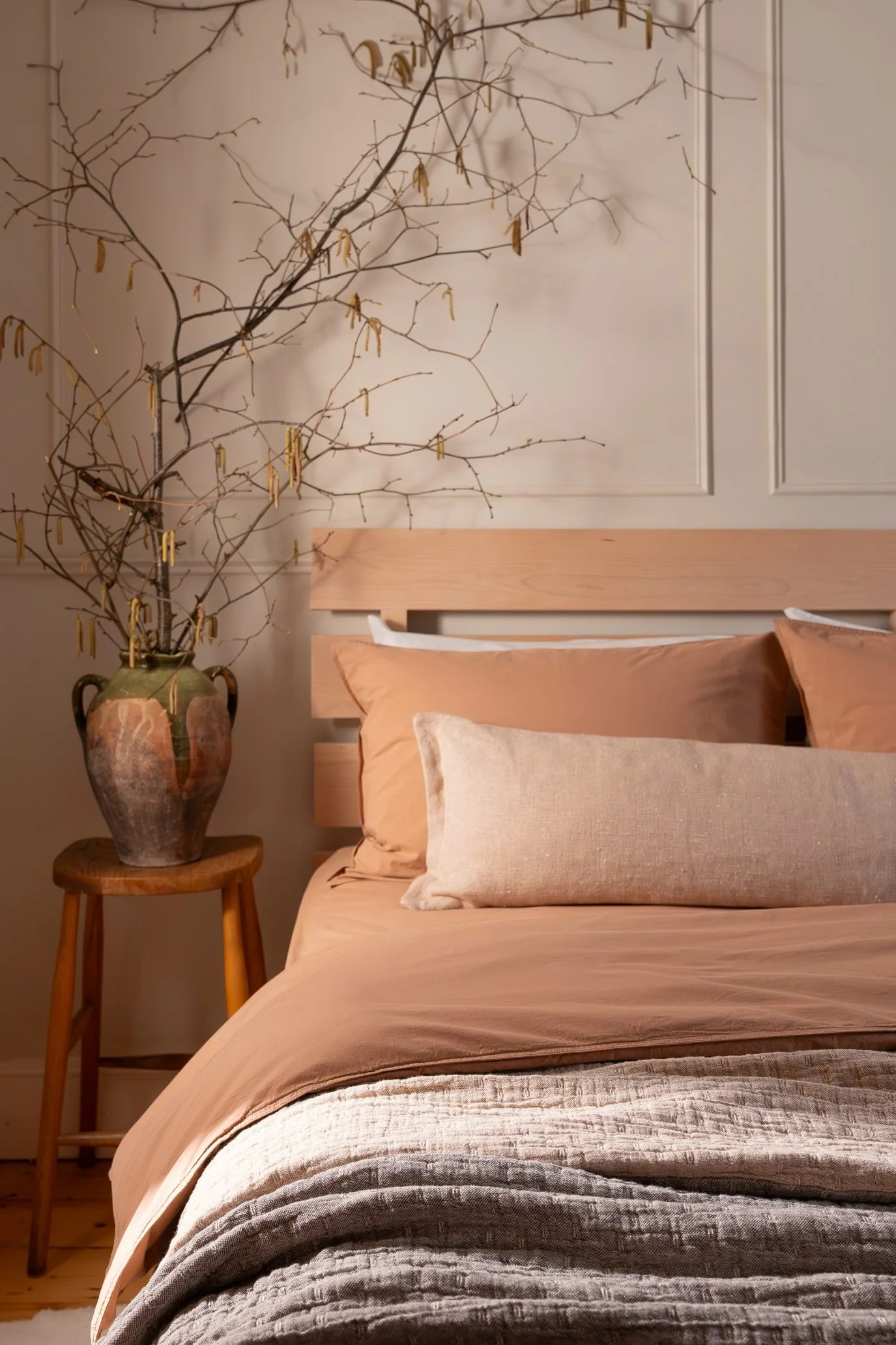 A styled bed in warm earthy tones with throws next to a side table with a vintage vase filled with branches