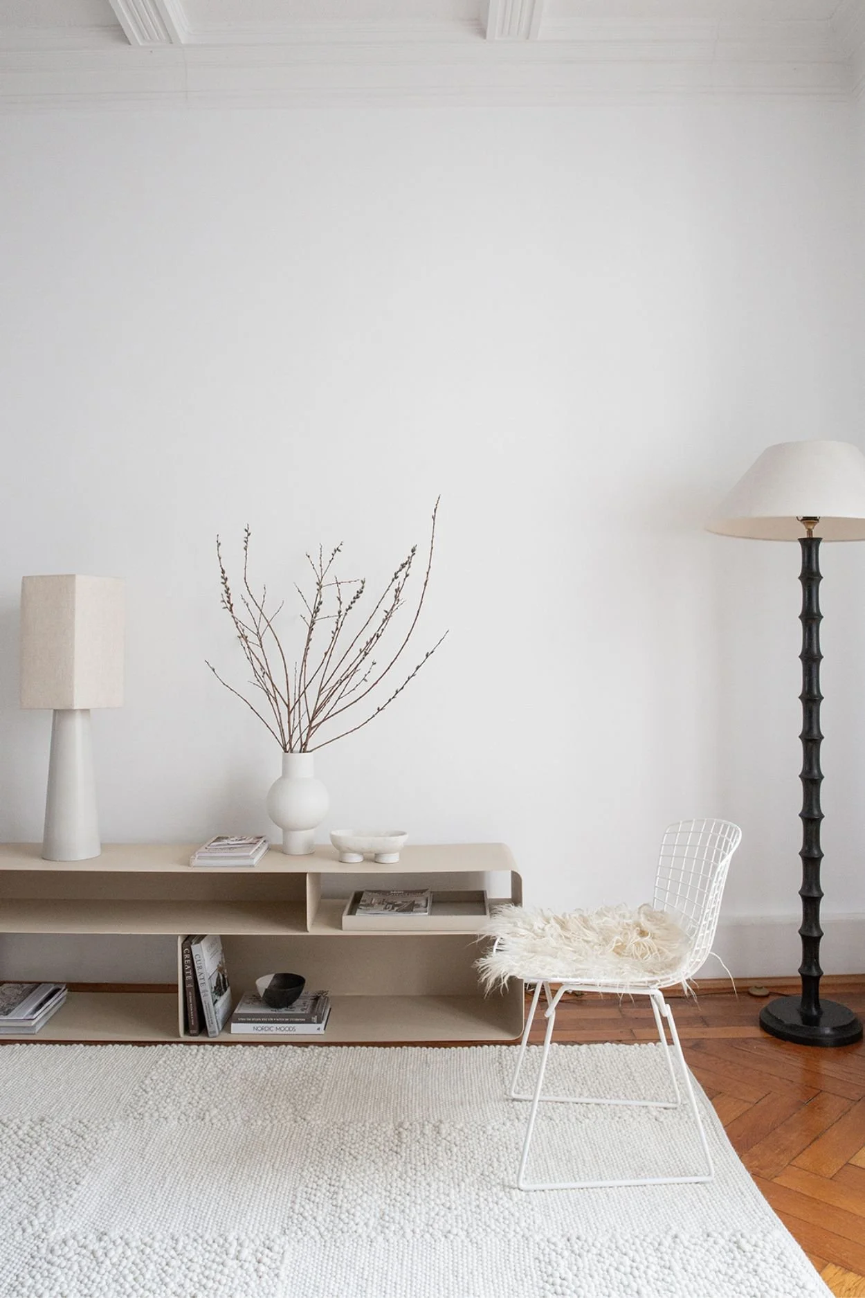 A room with a cream checkered wool rug and a floor lamp. On a unit there is a second lamp and vase in white