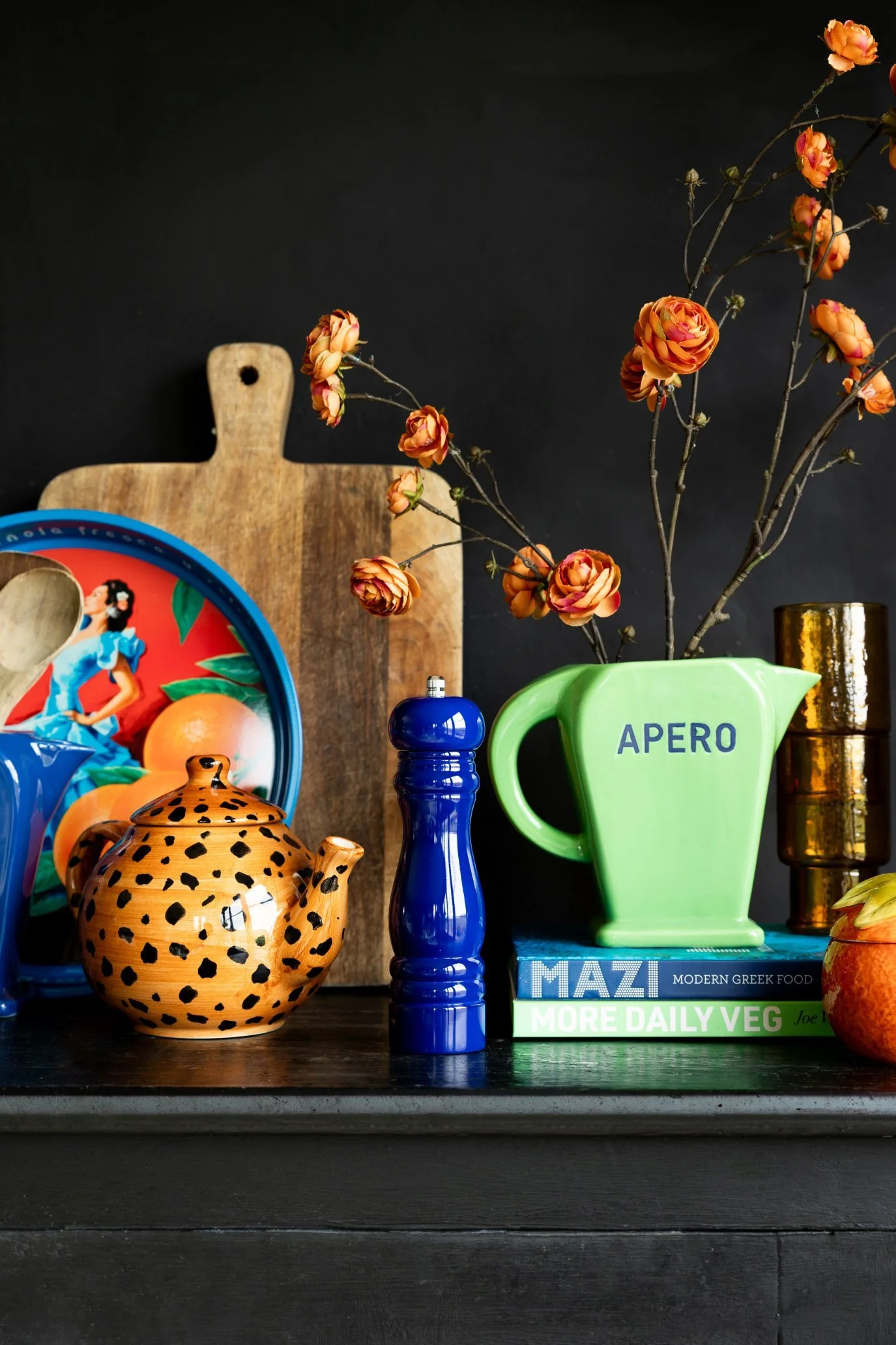 Bright coloured kitchen items like a jug, teapot and tray on a black shelf against a black wall