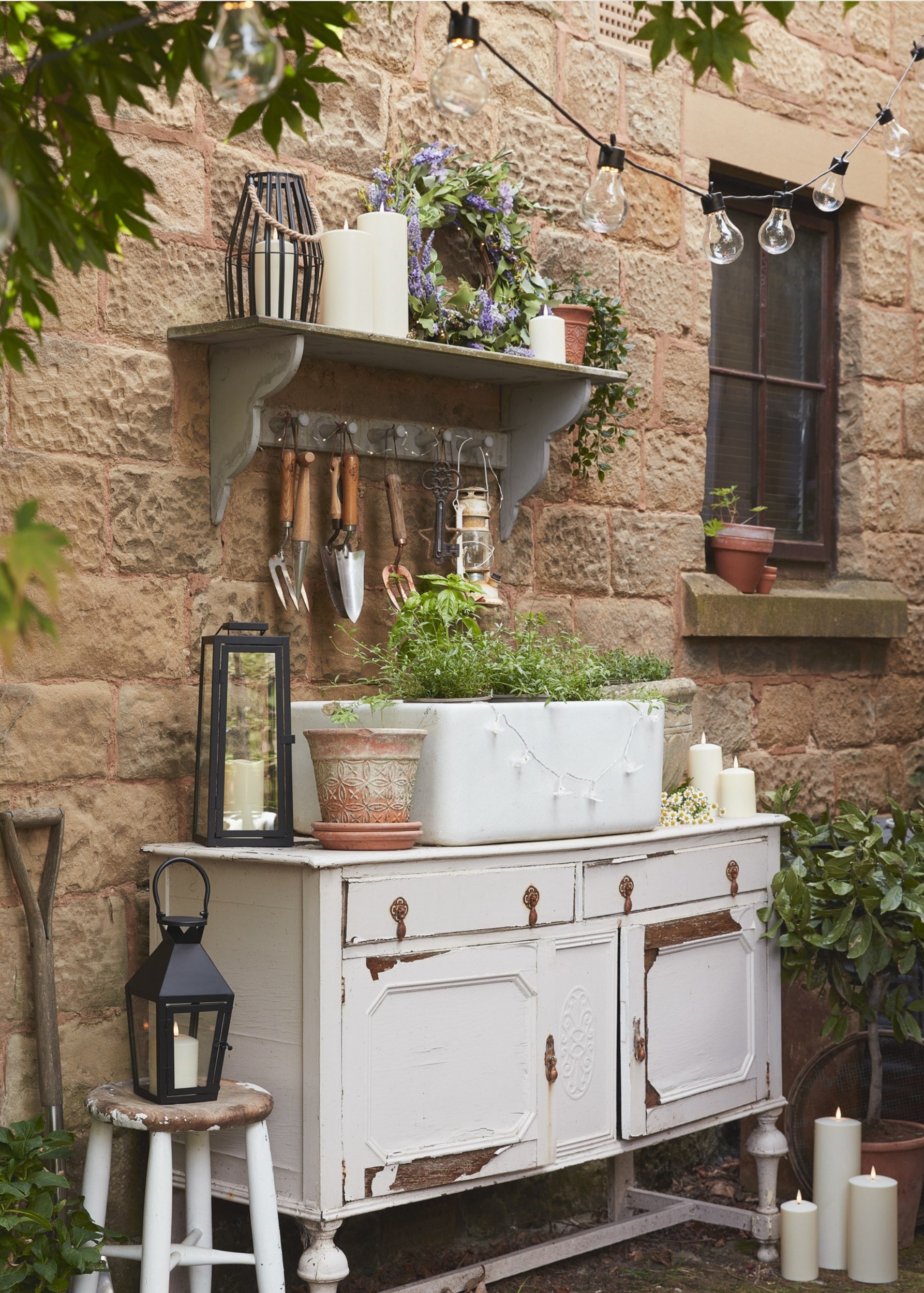 an old dresser in a garden with an old butlers sink full of herbs. The dresser and surrounding area are filled with lanterns and candles