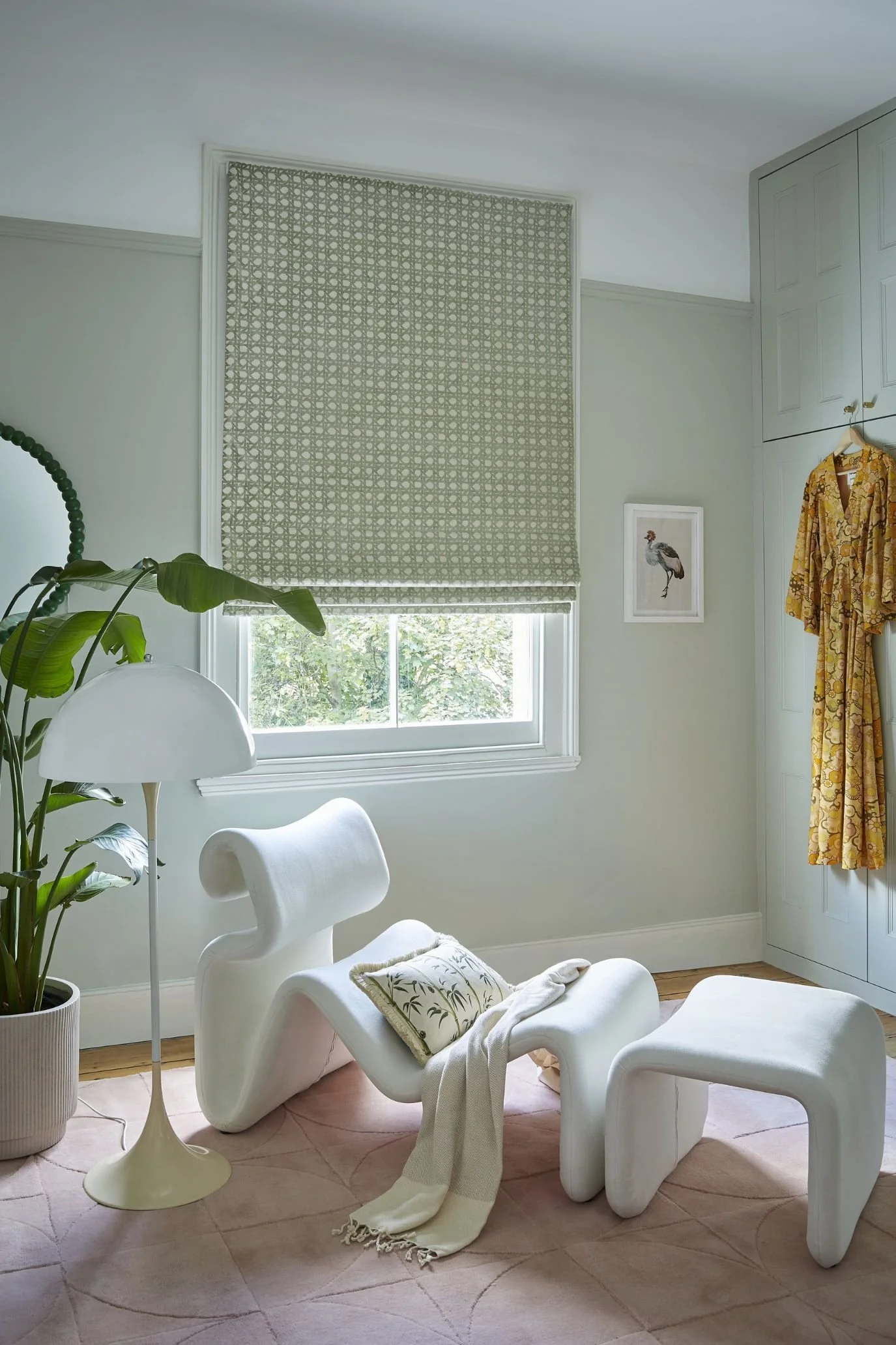 Soft green patterned Roman blind in a pale green room with sculptural white seating and a yellow dress hanging nearby.