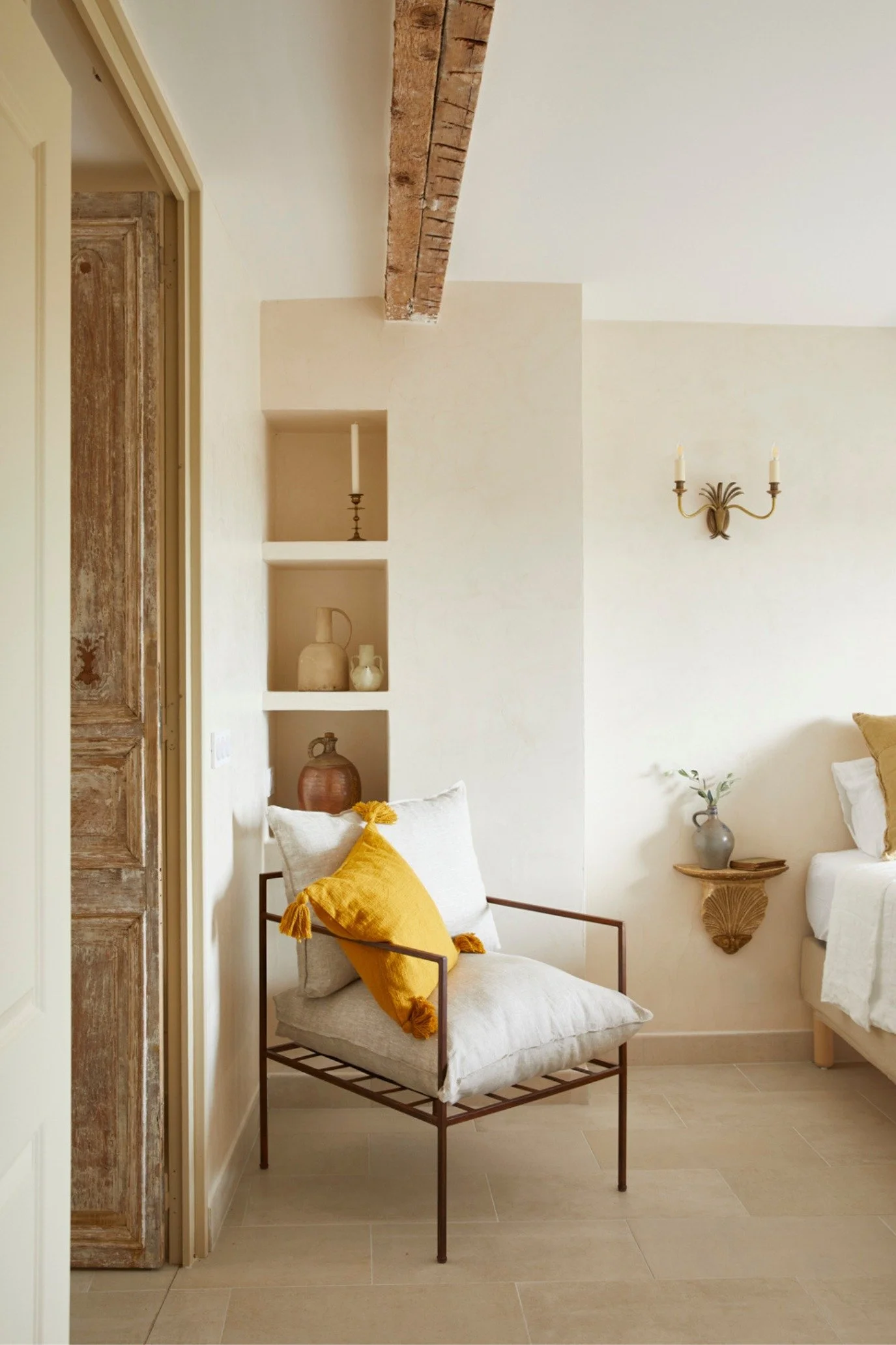 French provincial bedroom with limestone walls, exposed beams, ochre cushions, vintage iron chair and crisp white linen bedding.