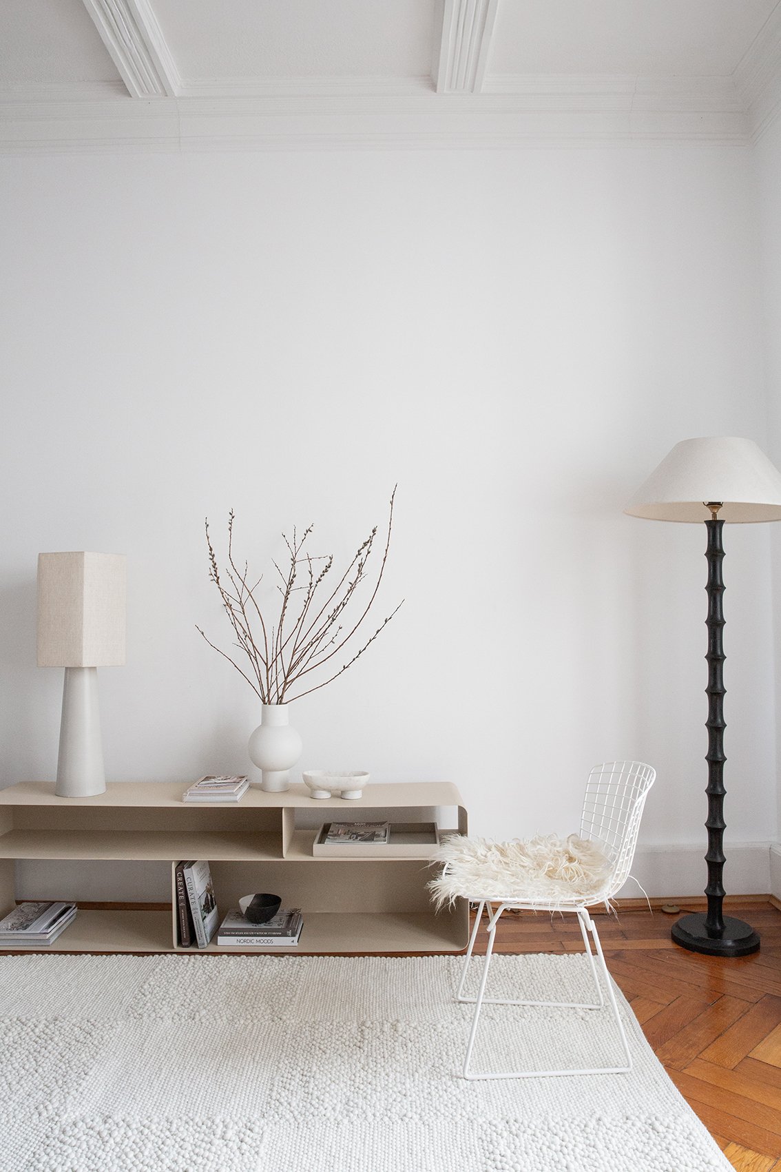 A room with a cream checkered wool rug and a floor lamp. On a unit there is a second lamp and vase in white