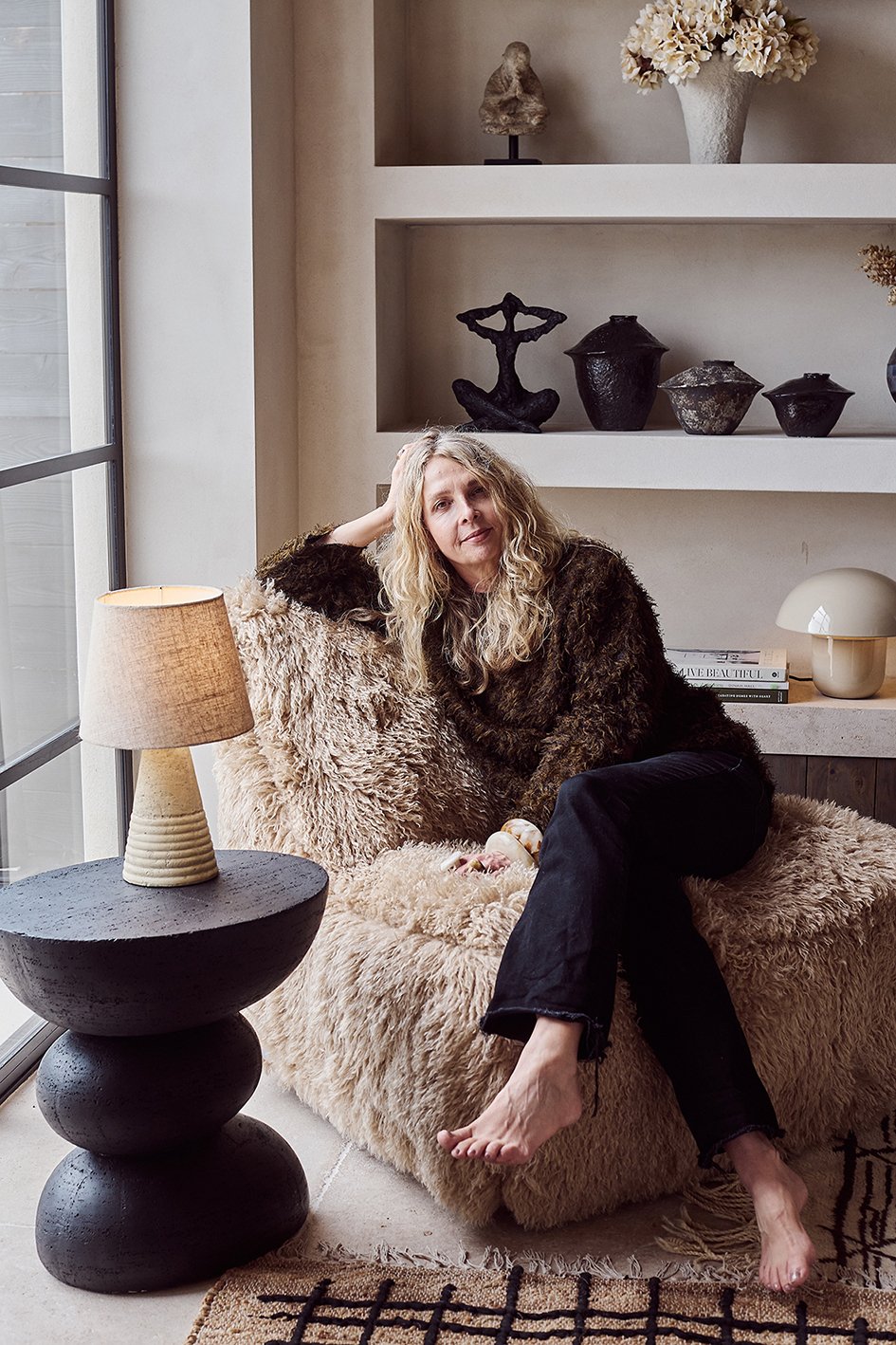 interior Designer Abigail Ahern sitting on a fur chair next to  a black round sculptured table with a cream lamp on it with recessed shelves behind
