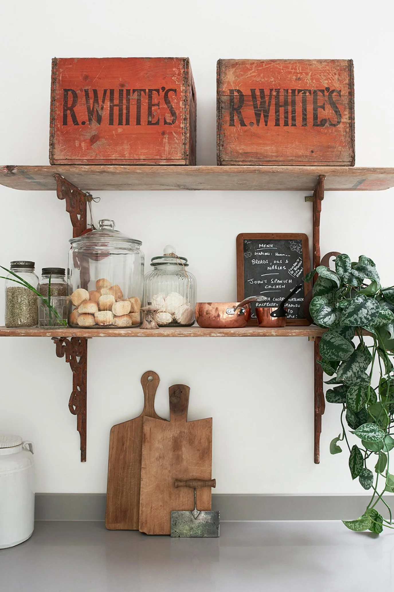 Vintage brackets with aged wooden shelves with two vintage r.whites boxes on top and decorative kitchen items like jars, copper pans next to a trailing plant and min blackboard