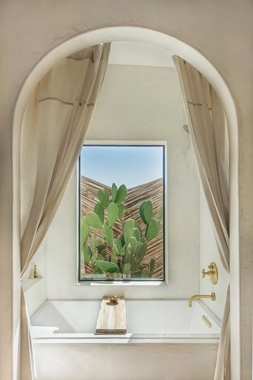 deep soaking tub sitting below a plaster arch with curtains pulled back to one side and a window looking over a cactus