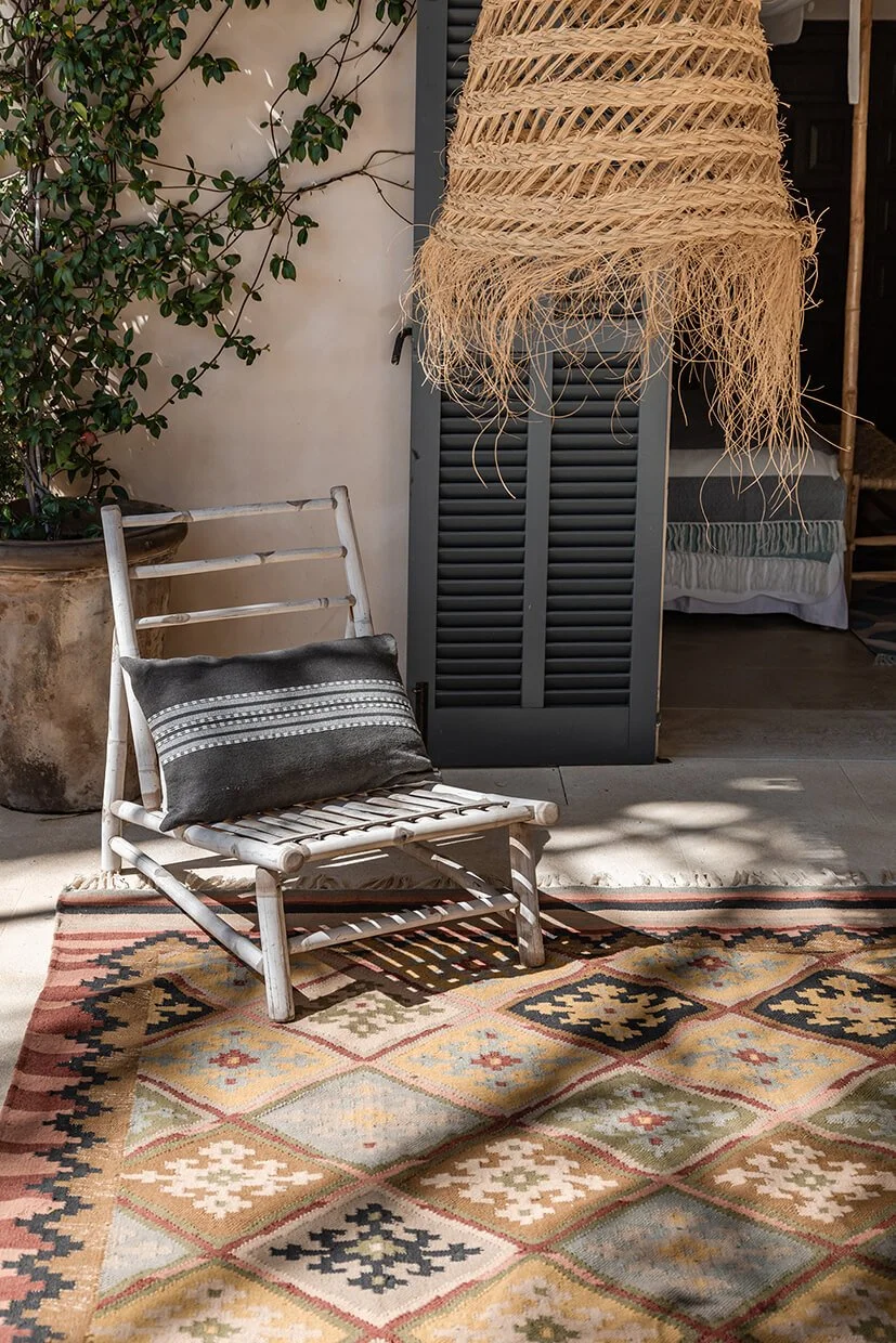White bamboo chair with a dark grey striped cushion on a patterned outdoor rug by an open terrace doorway