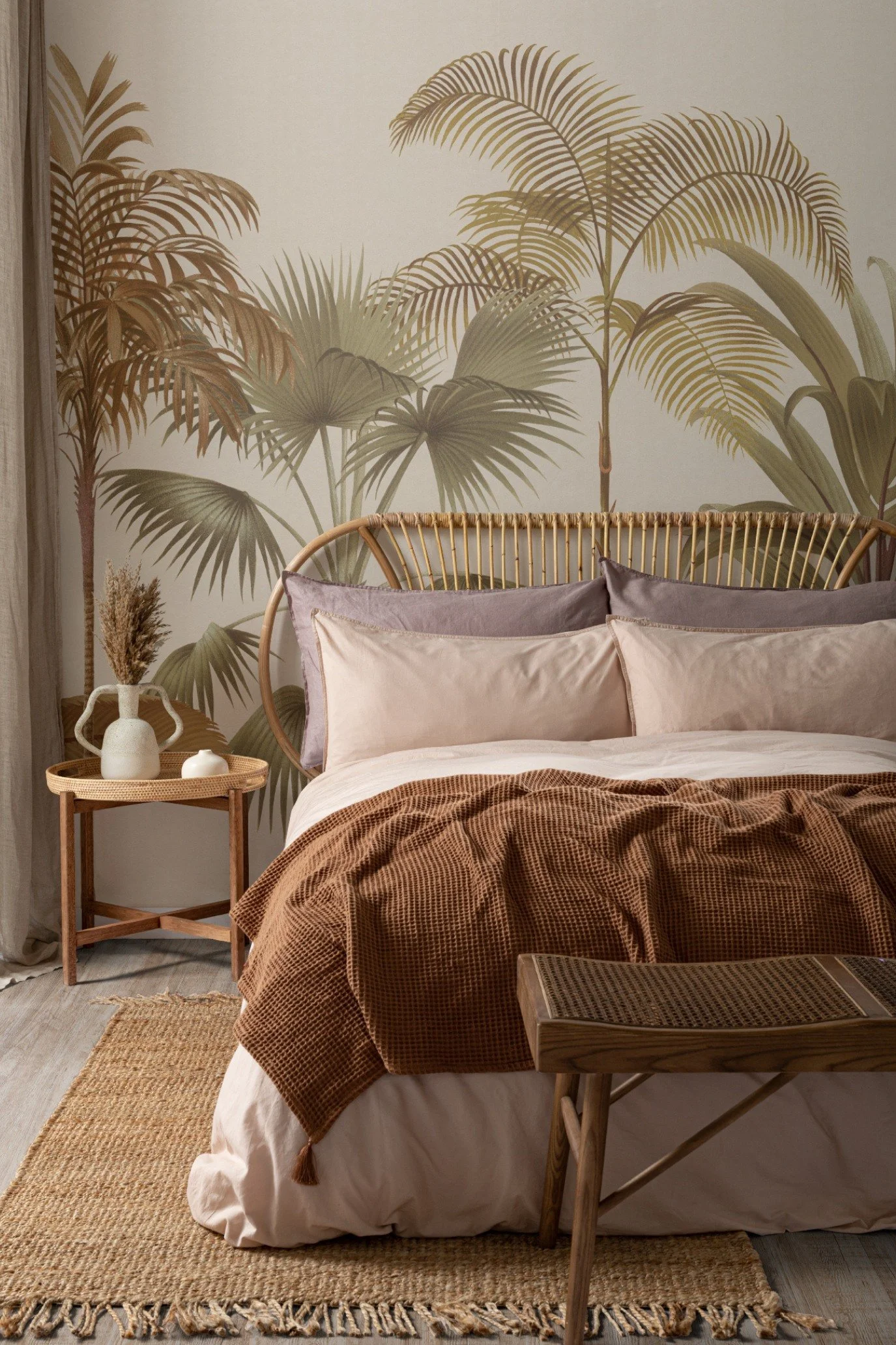 A bed in front of a wall with a palm tree mural with arattan beside table and stool at the end of the bed which has a bamboo headboard