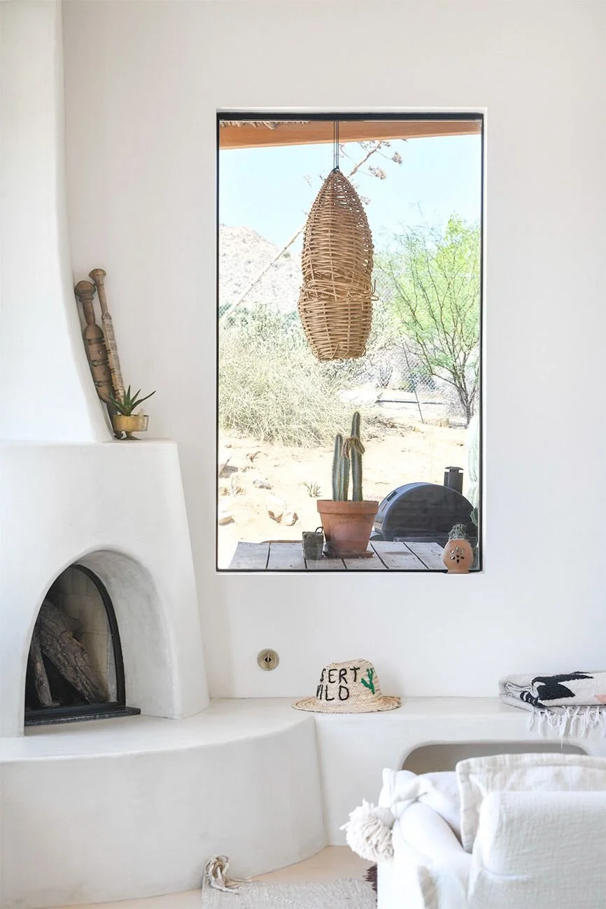 A Georgia O’Keeffe–inspired adobe fireplace next to a window looking out at the terrace table with a potted cactus and rattan pendant light
