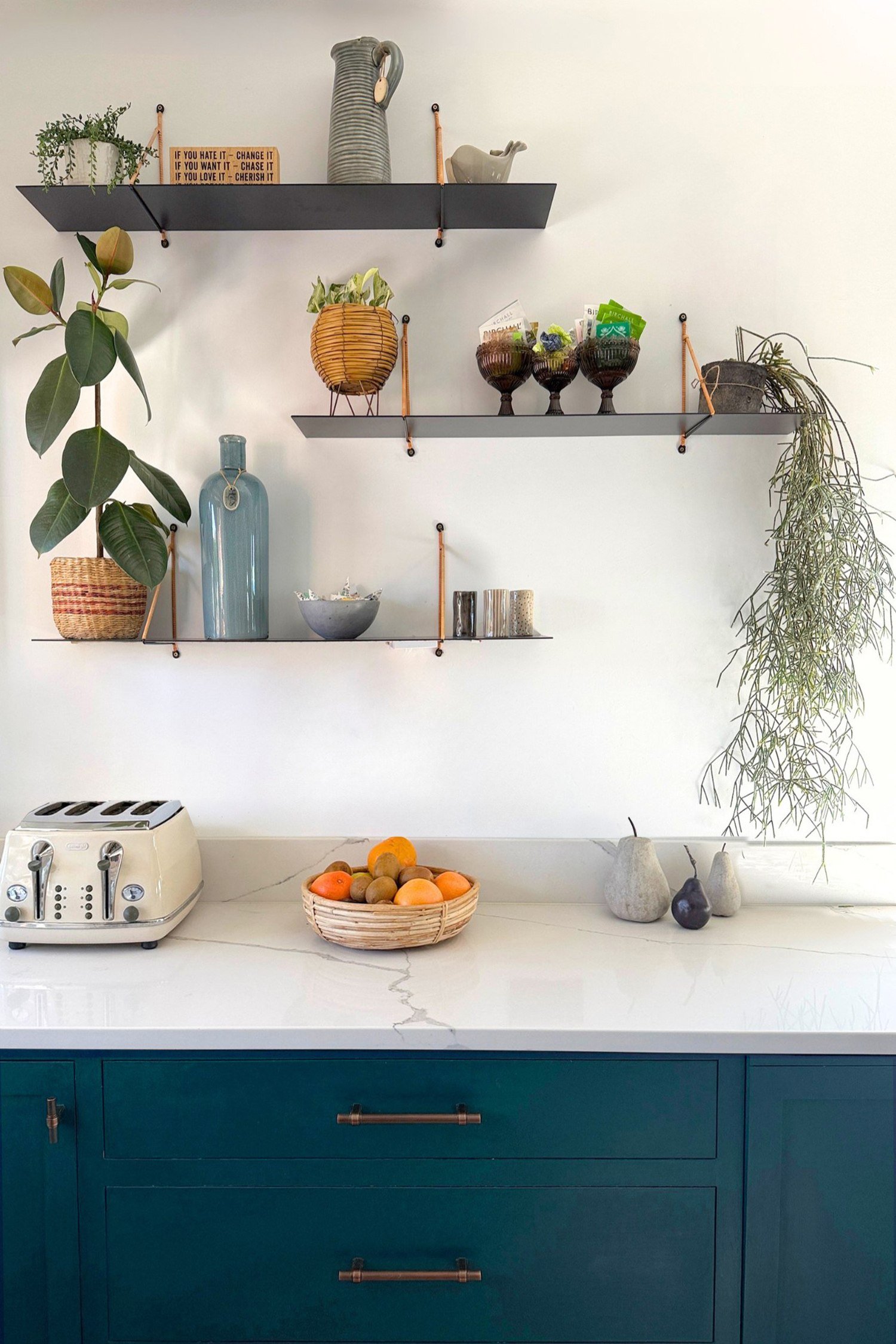 A kitchen in teal cabinetry with a white marble countertop and three black metal shelves styled with plants and ceramic vessels