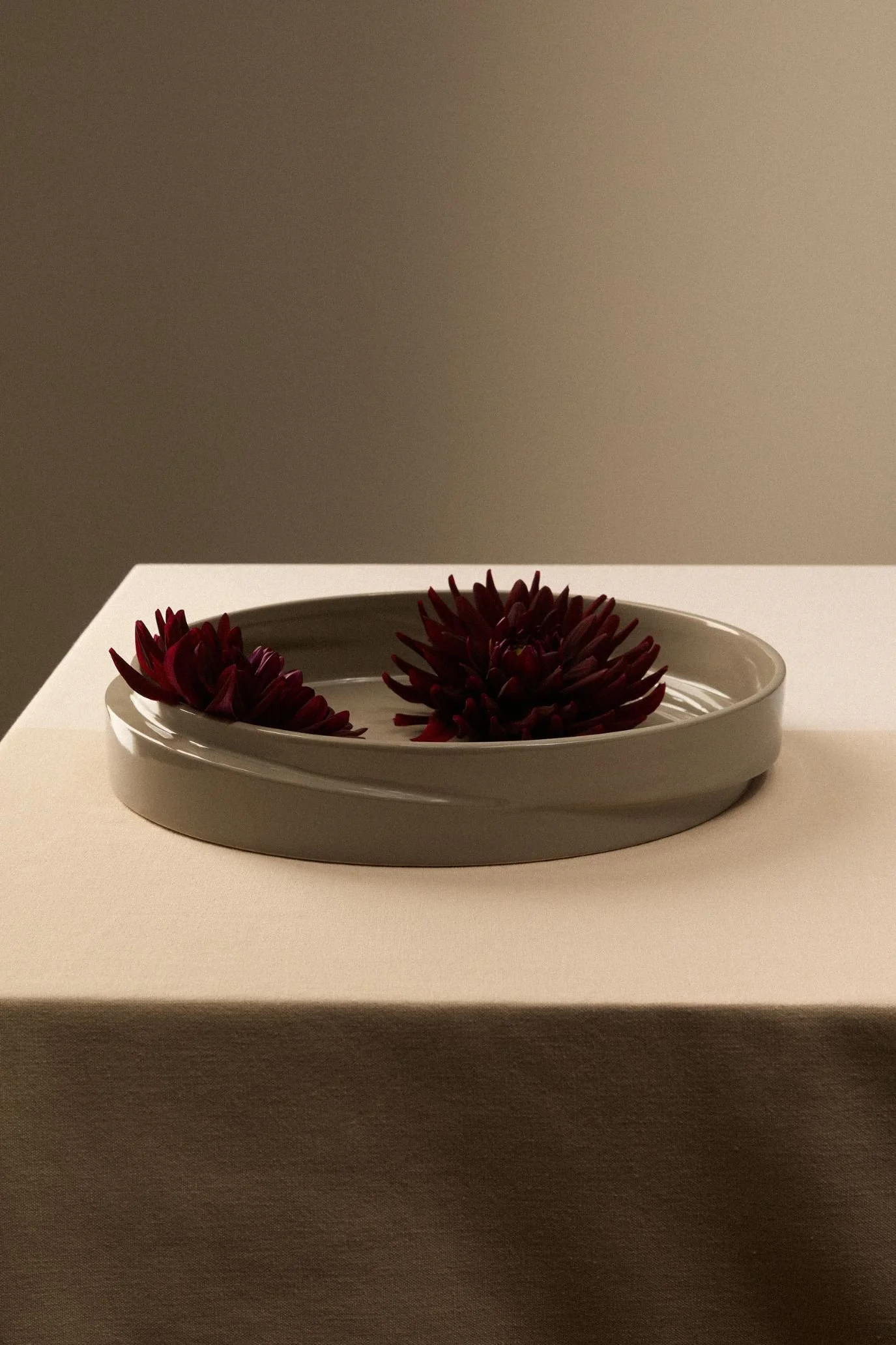 Kelly Hoppen x M&S shallow neutral bowl holding dark burgundy decorative stems on a fabric-covered table.