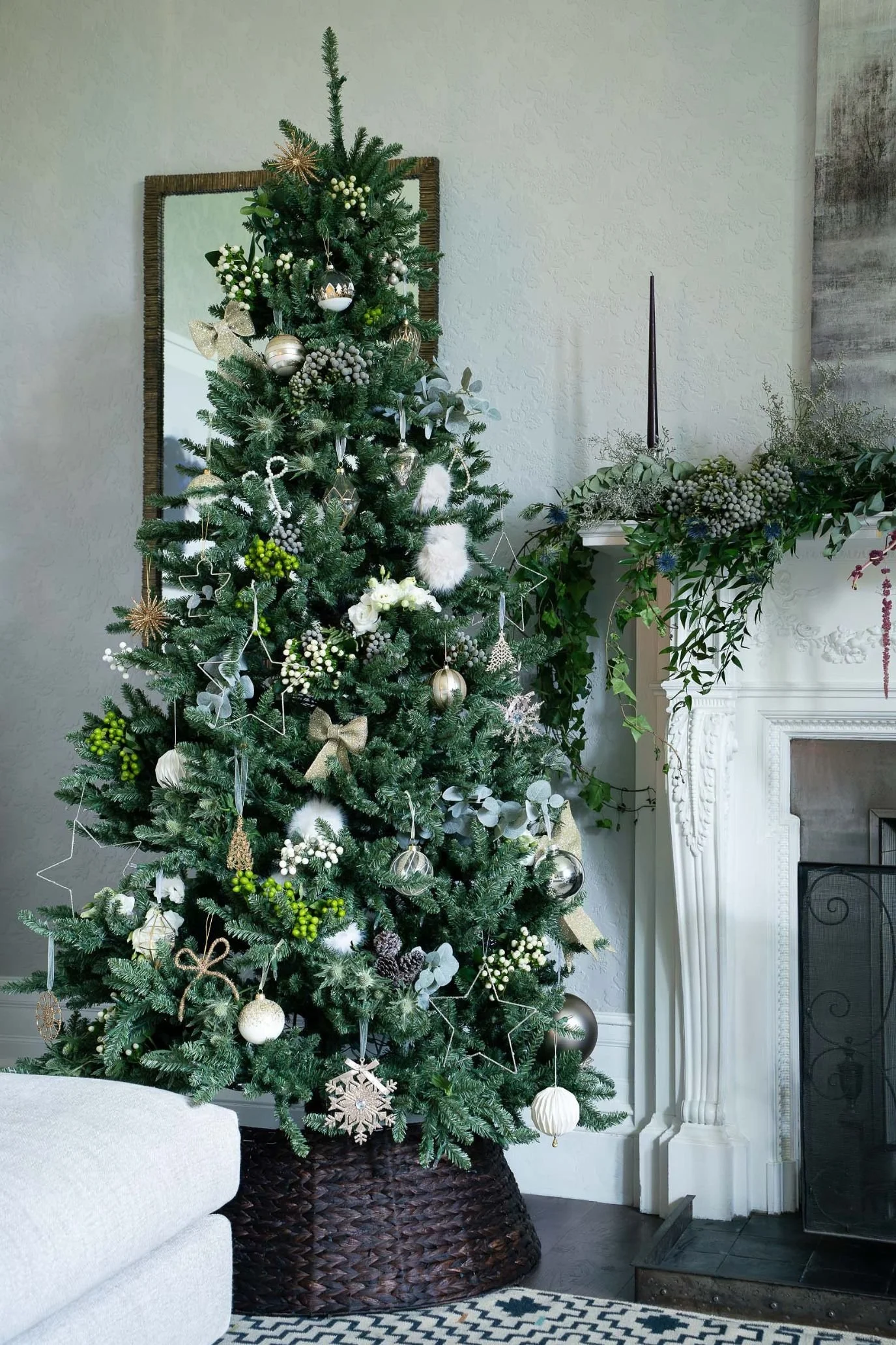 Christmas tree in a dark woven basket, decorated with small white flower bunches, greenery and pale hanging ornaments.