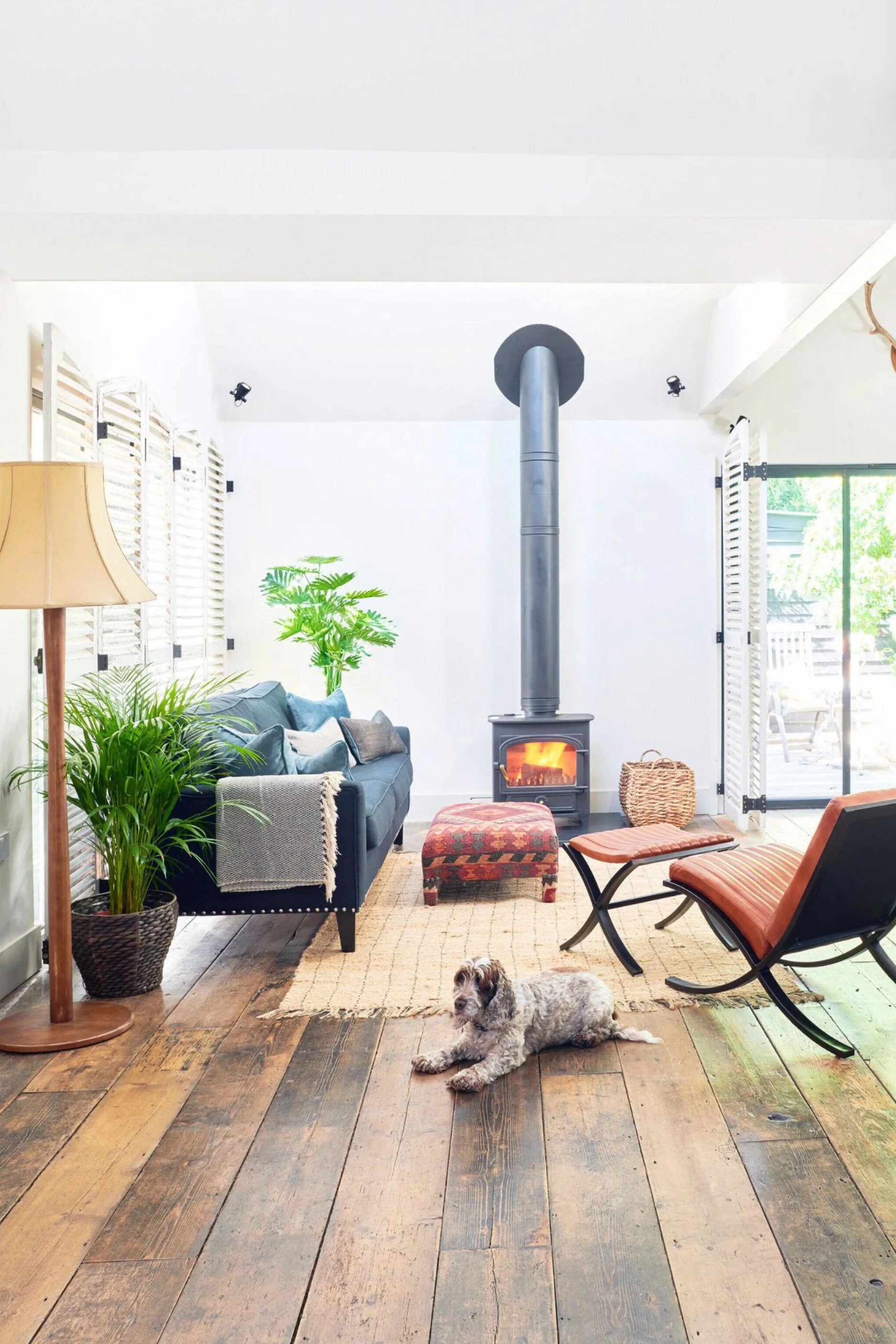 A living room with a wood burning stove alight with a blue sofa infront and a red ottoman. Towards the front a spaniel is on the floor close to a tall wood lamp with a white shade