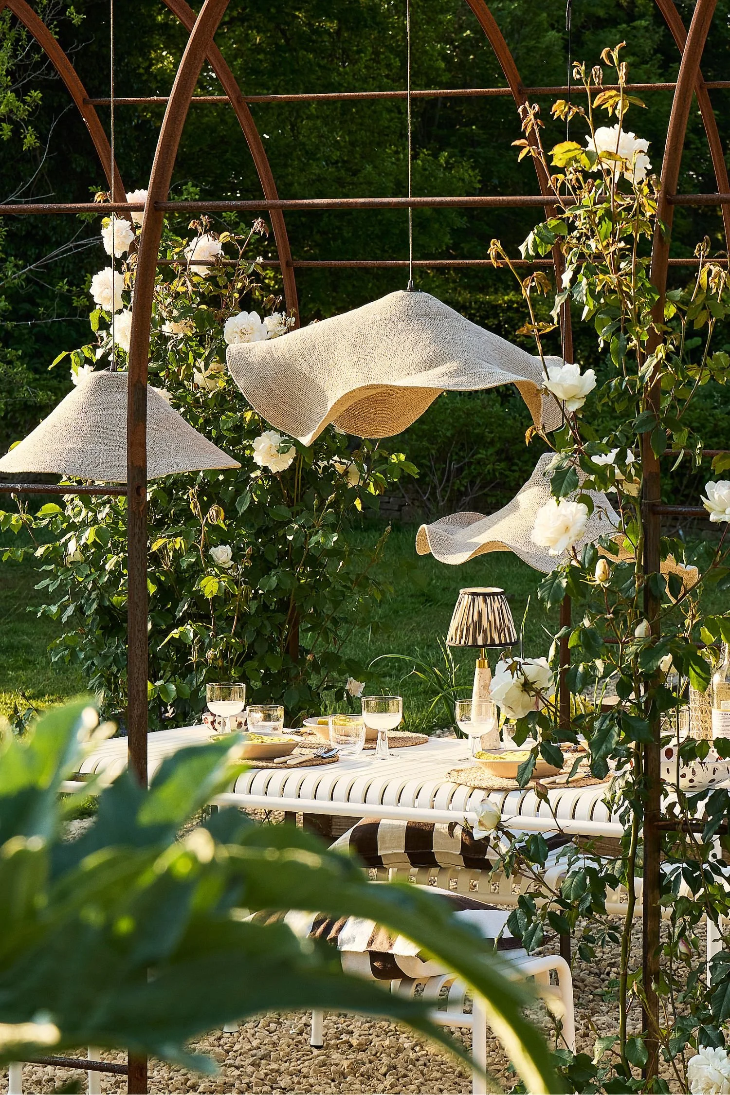 Outdoor lighting ideas featuring sculptural woven pendant lights above a stylish outdoor dining area covered with climbing roses.