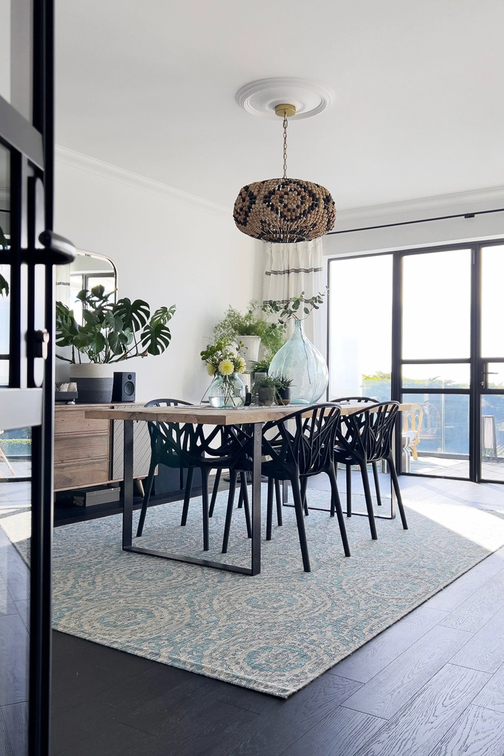 Dining room with blue rug a beaded pendant light and plants on surfaces looking out to black framed doors