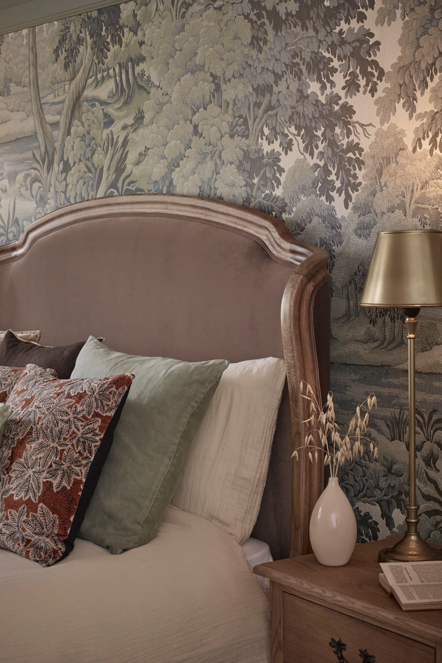 A carved headboard with mauve velvet against a wallpaper with trees. There are green and pink cushions on the bed and a lamp and white vase next to the bed