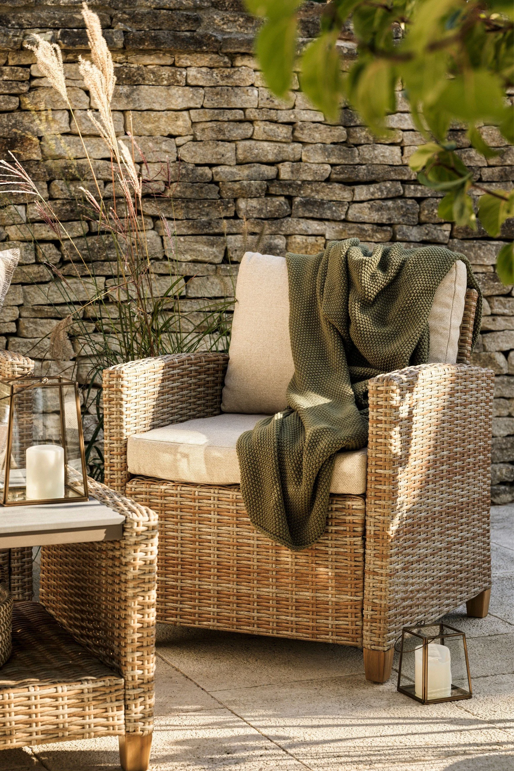 an outdoor wicker chair with a green throw on it next to a low rattan coffee table with a lantern on it