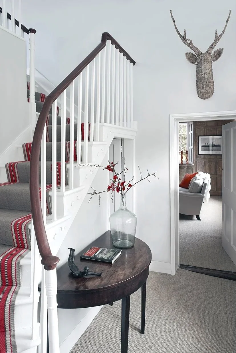 A staircase with a beige and red edged runner with an antique console table infront with flowers and a book  and a rattan deer head