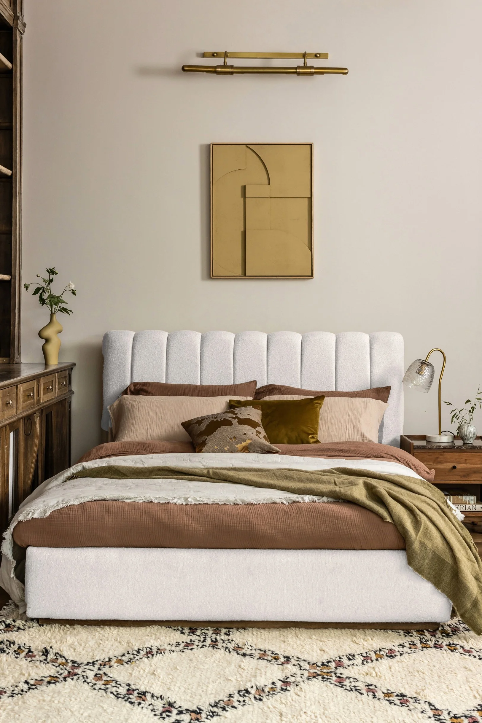 A bedroom with a velvet fluted off-white headboard and a bed dressed in brown green and white bedlinen. There is a picture above the bed with a light fixture above it