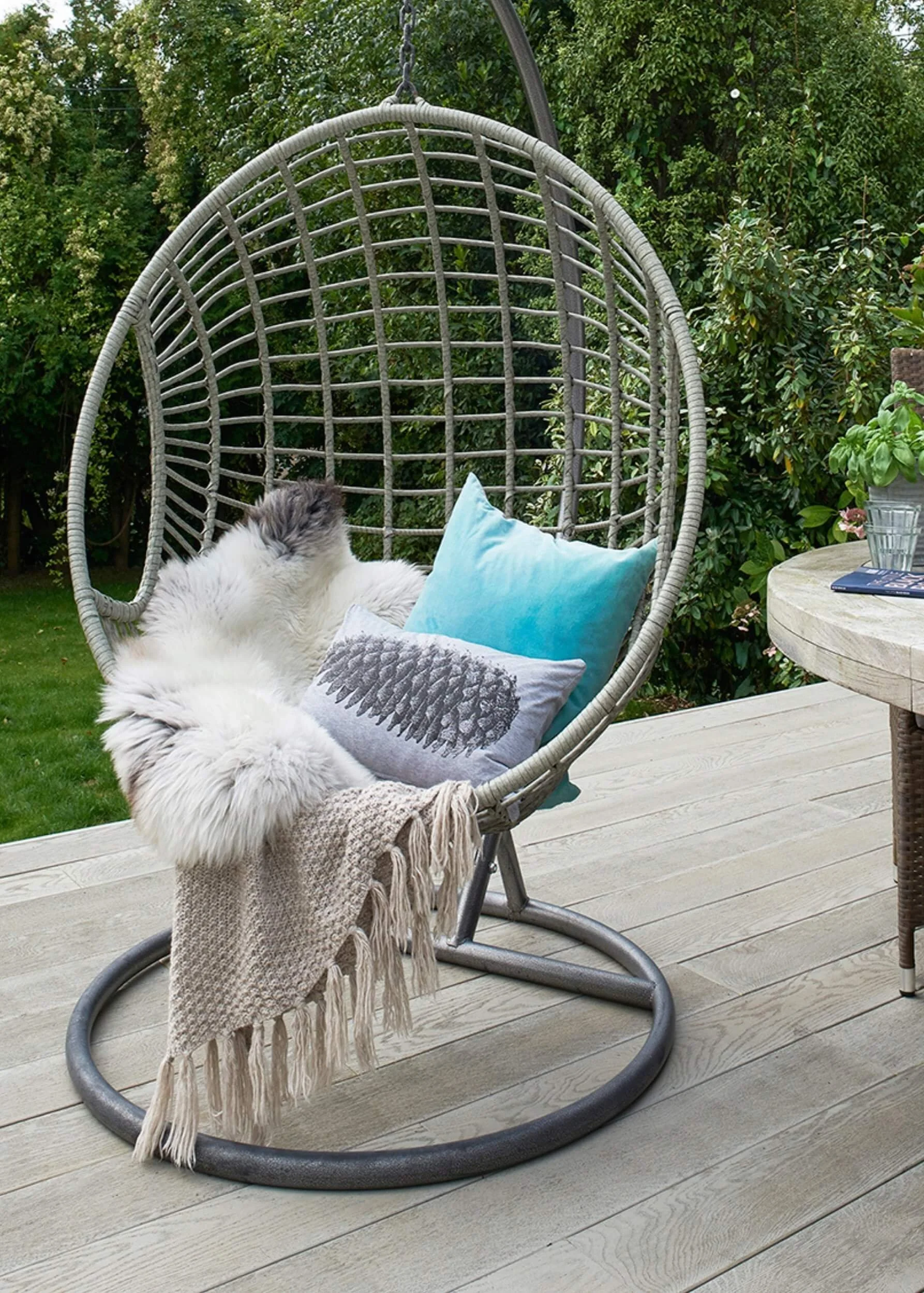 a freestanding egg swing chair styled with two cushions, a sheepskin and a hassled throw in a garden on a decking