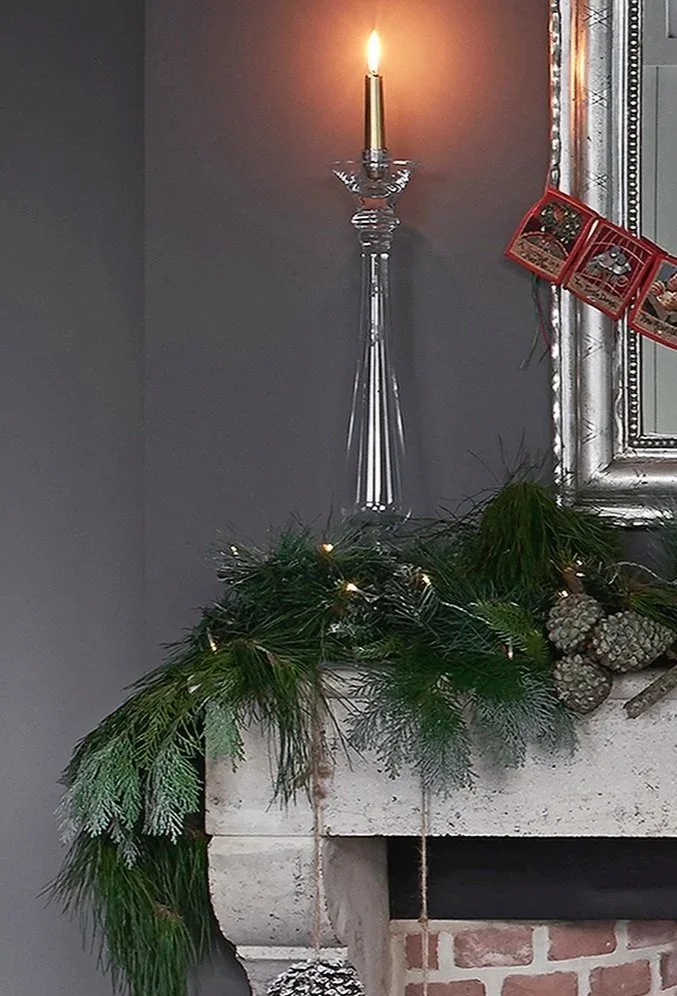 A glass candle holder with a gold candle lit on a rustic mantlepiece covered in fir and pinecones